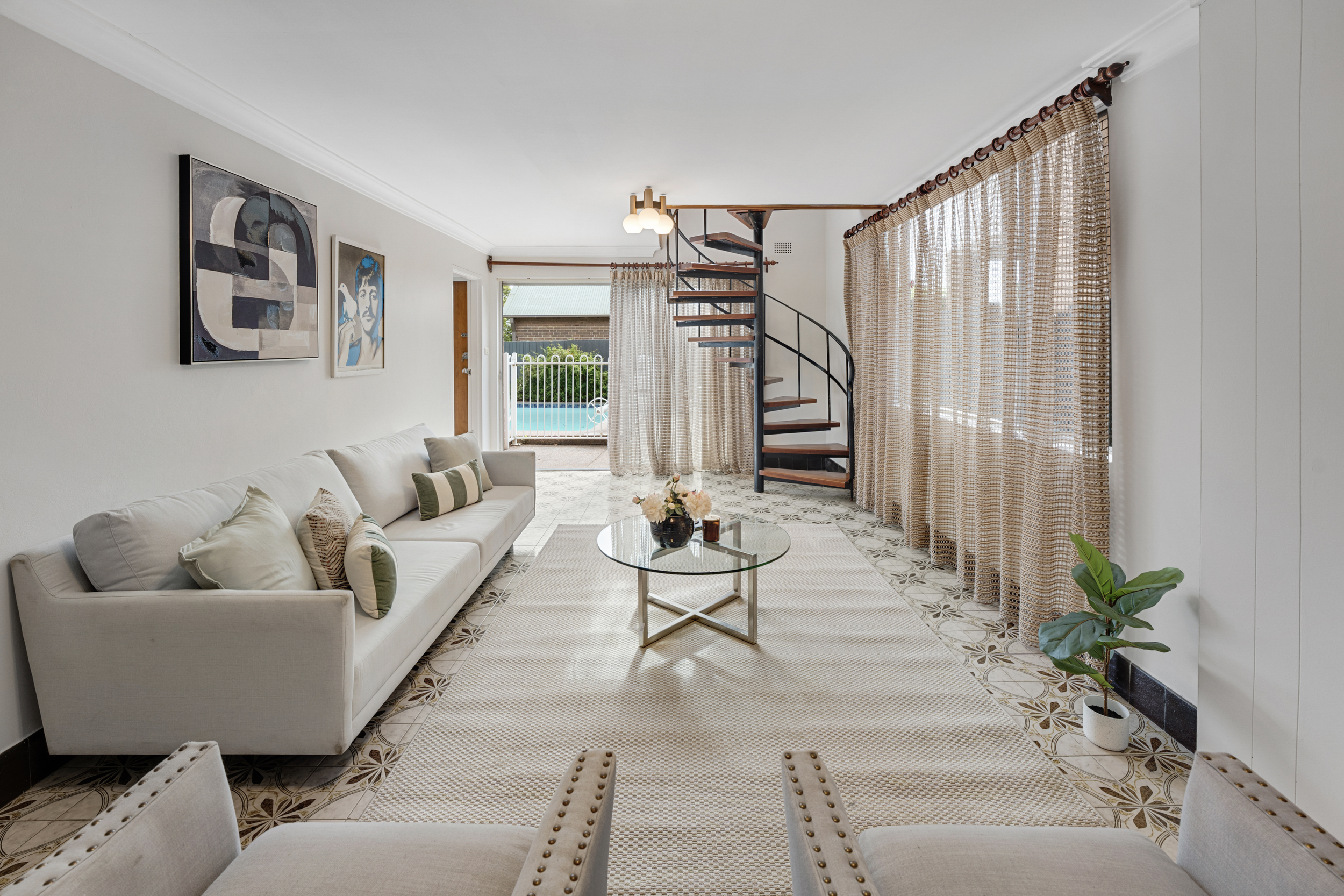 Living room with white sofa, patterned tile floor, glass coffee table with flowers, window with curtains, spiral staircase, wall art, potted plant.