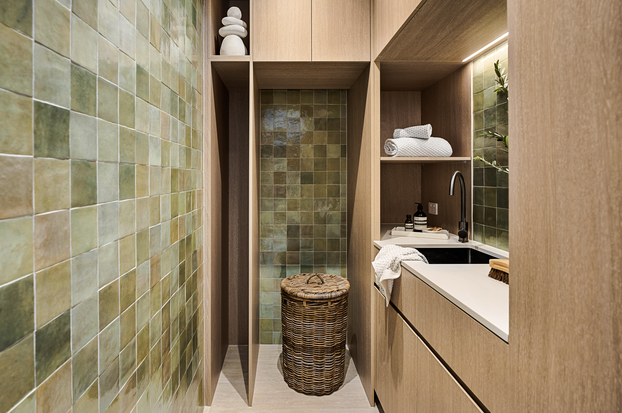 A small modern bathroom with green tile walls, wooden cabinets, a wicker laundry basket, folded towels, and a black sink.