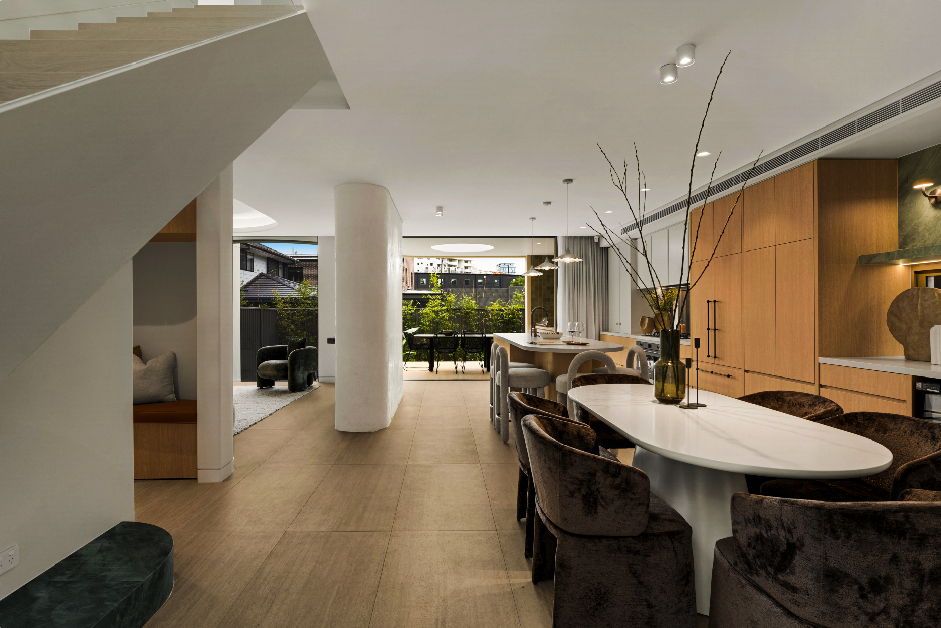 Modern open-concept living space with dining area and kitchen, featuring a white oval table with velvet chairs, wooden cabinetry, and decorative vases with bare branches, overlooking an outdoor patio with greenery.