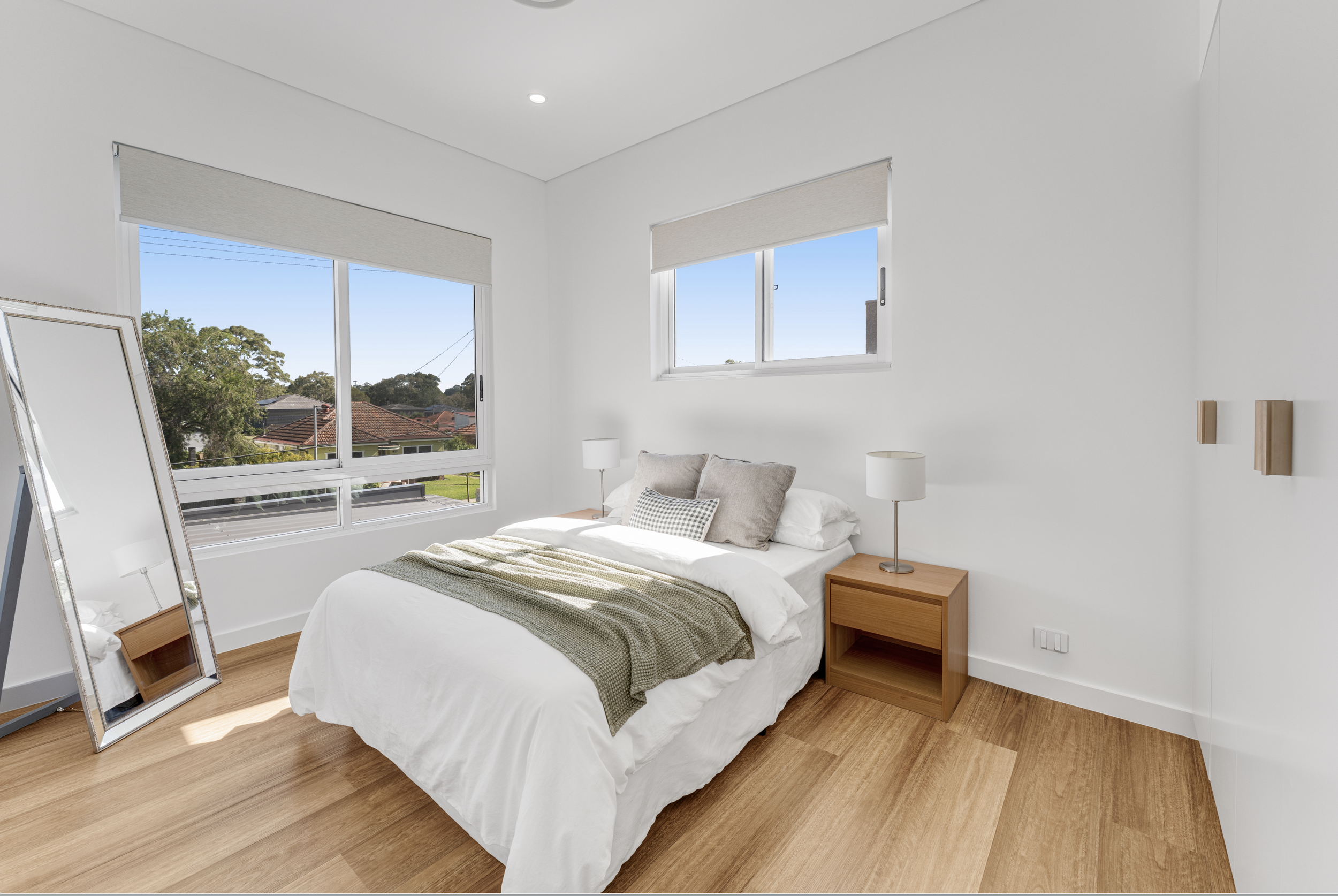 A bedroom with white walls, two large windows with blinds, a bed with white linens, a beige throw blanket, and pillows. There are two wooden nightstands with lamps, and a full-length mirror leaning against the wall. The room has a hardwood floor.