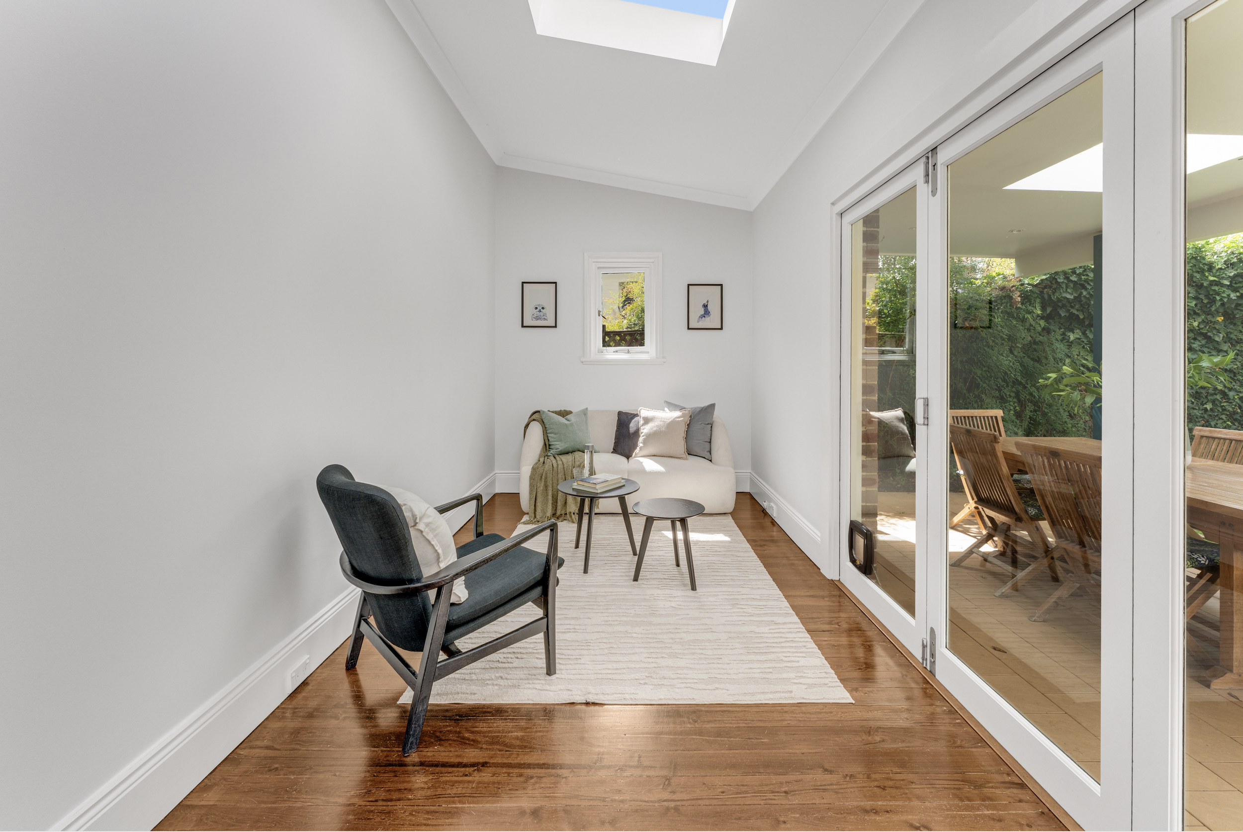 A bright living room with white walls and ceiling, hardwood floors, large glass sliding door leading to an outdoor patio with wooden chairs, a sofa with cushions, and small coffee tables. Skylights in the ceiling provide natural light.