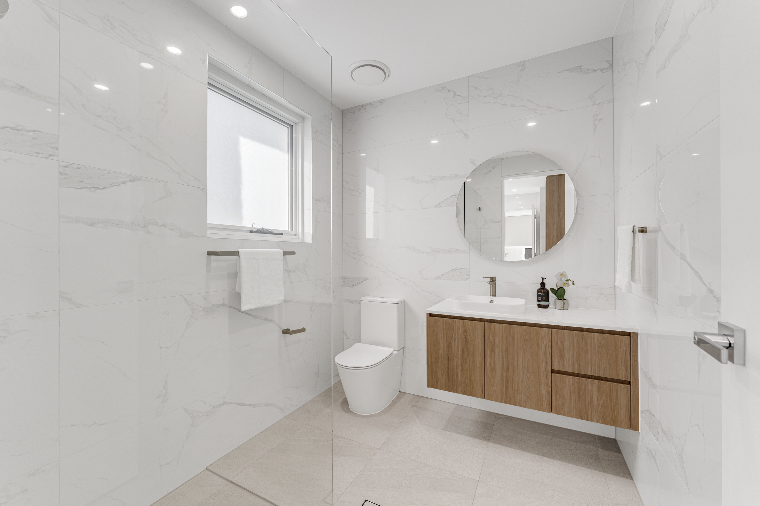 Modern bathroom with white marble walls and floor, a window with a frosted glass panel, a round mirror above a wooden vanity with sink, towel rack with white towels, toilet, and decorative plants.