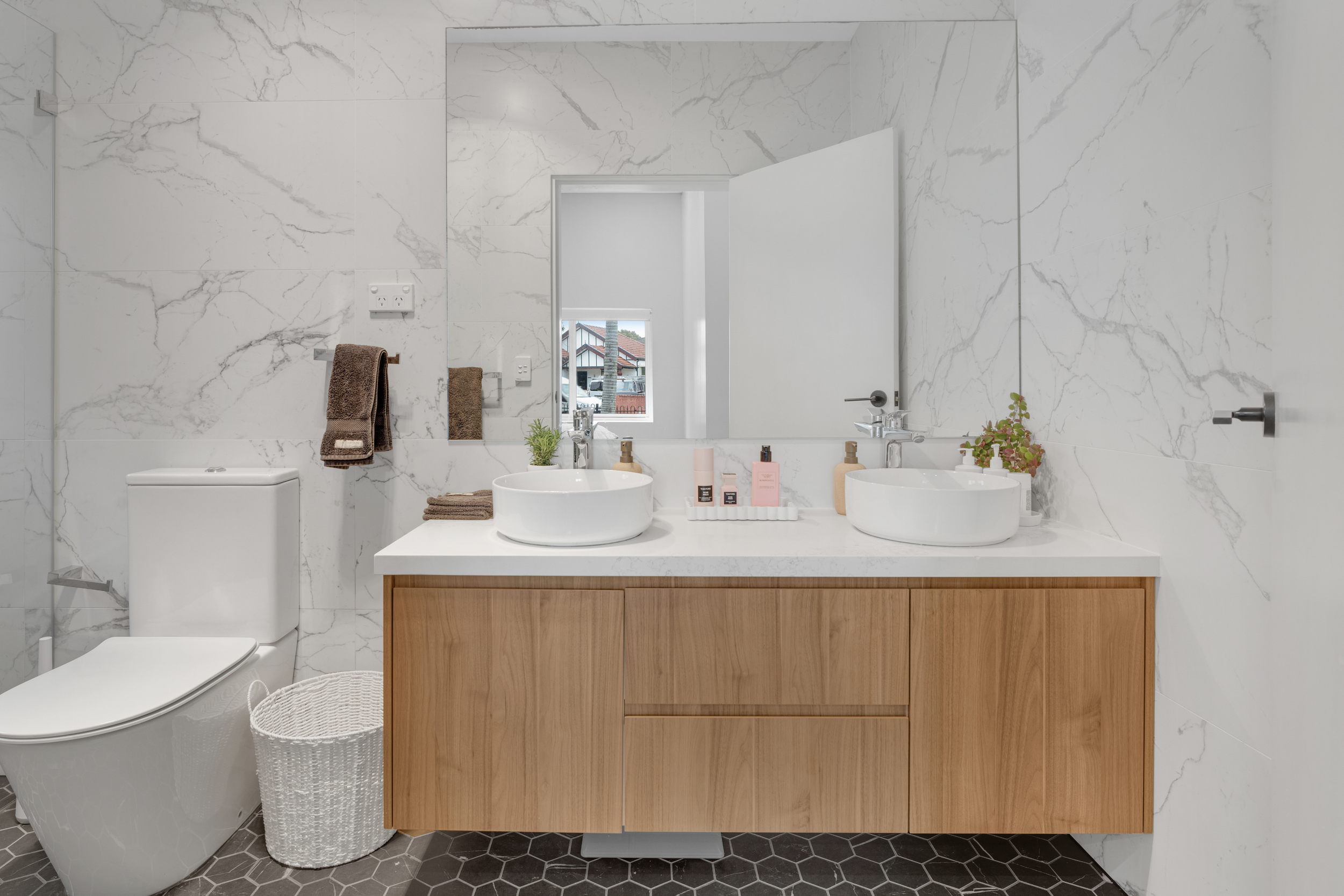 Modern bathroom with white marble walls, a wooden vanity with two vessel sinks, a large mirror, and a window providing natural light. Decor includes small plants, soap, and lotion bottles. There is a white toilet, brown towels, a trunk-style laundry 
