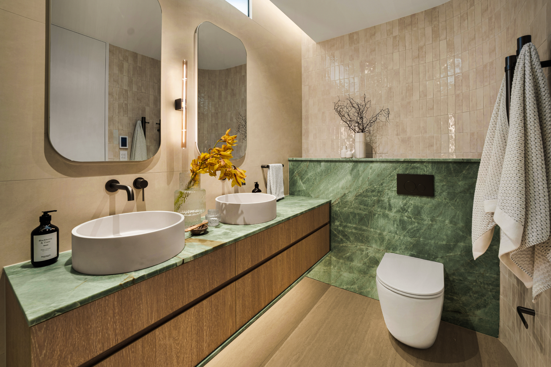 Modern bathroom with double oval vessel sinks on a green marble countertop, framed by large mirrors, wooden cabinetry, and beige tiled walls. Decor includes vases with flowers and branches, towel rack with textiles, and a wall-mounted toilet.