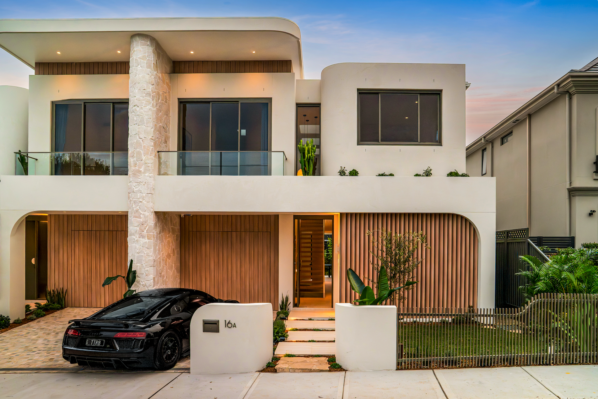 Modern two-story house with a beige exterior, large windows, and wooden accents. The house has a white wall, a small front garden, a gated entrance, and a black luxury sports car parked in the driveway. The sky is partly cloudy, and there are plants 