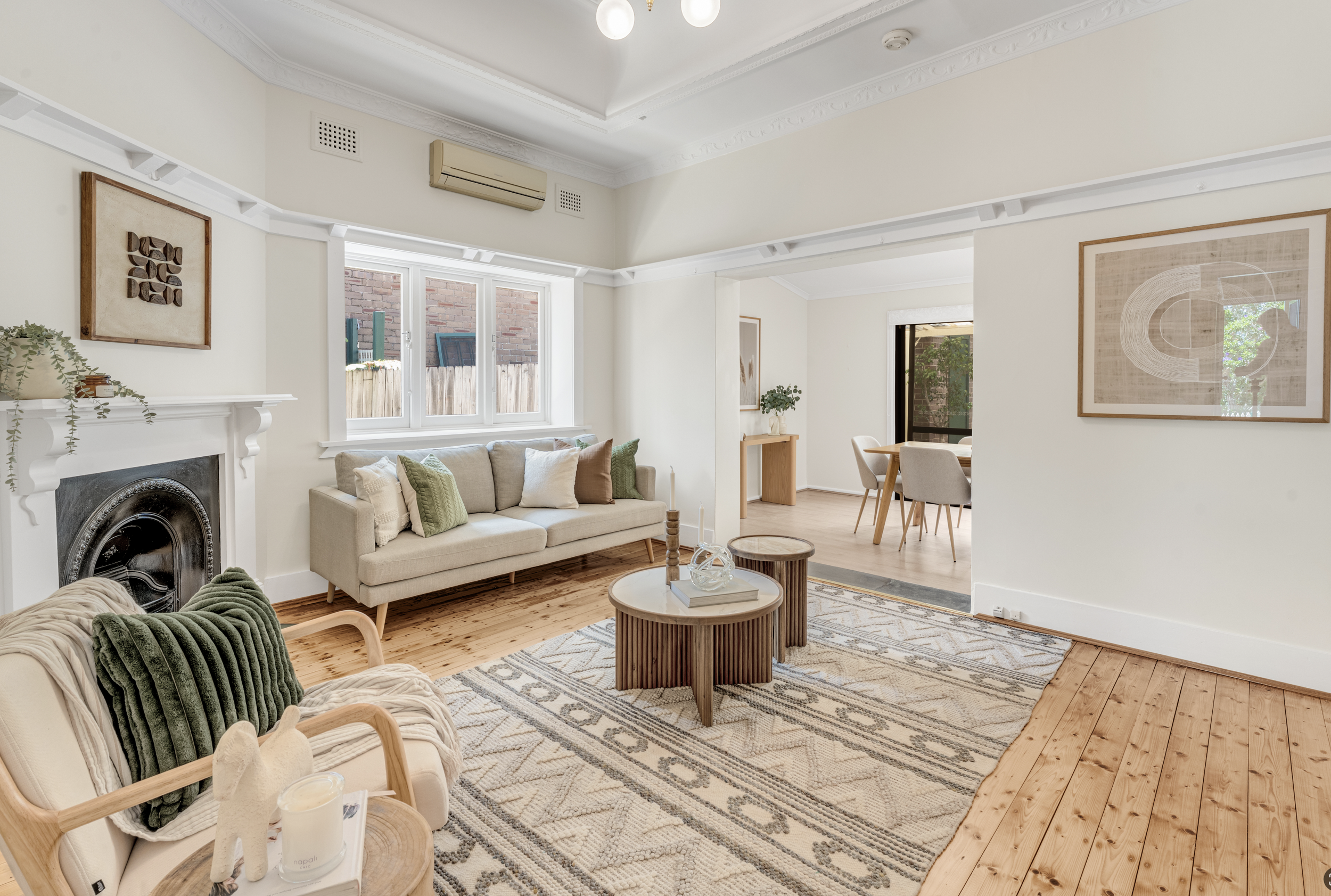 A living room with a cream-colored sofa, a beige armchair with a green throw blanket, a glass coffee table with books and decor, a patterned area rug, a fireplace with a mantel and plant, large window showing a brick wall outside, and artwork on the 