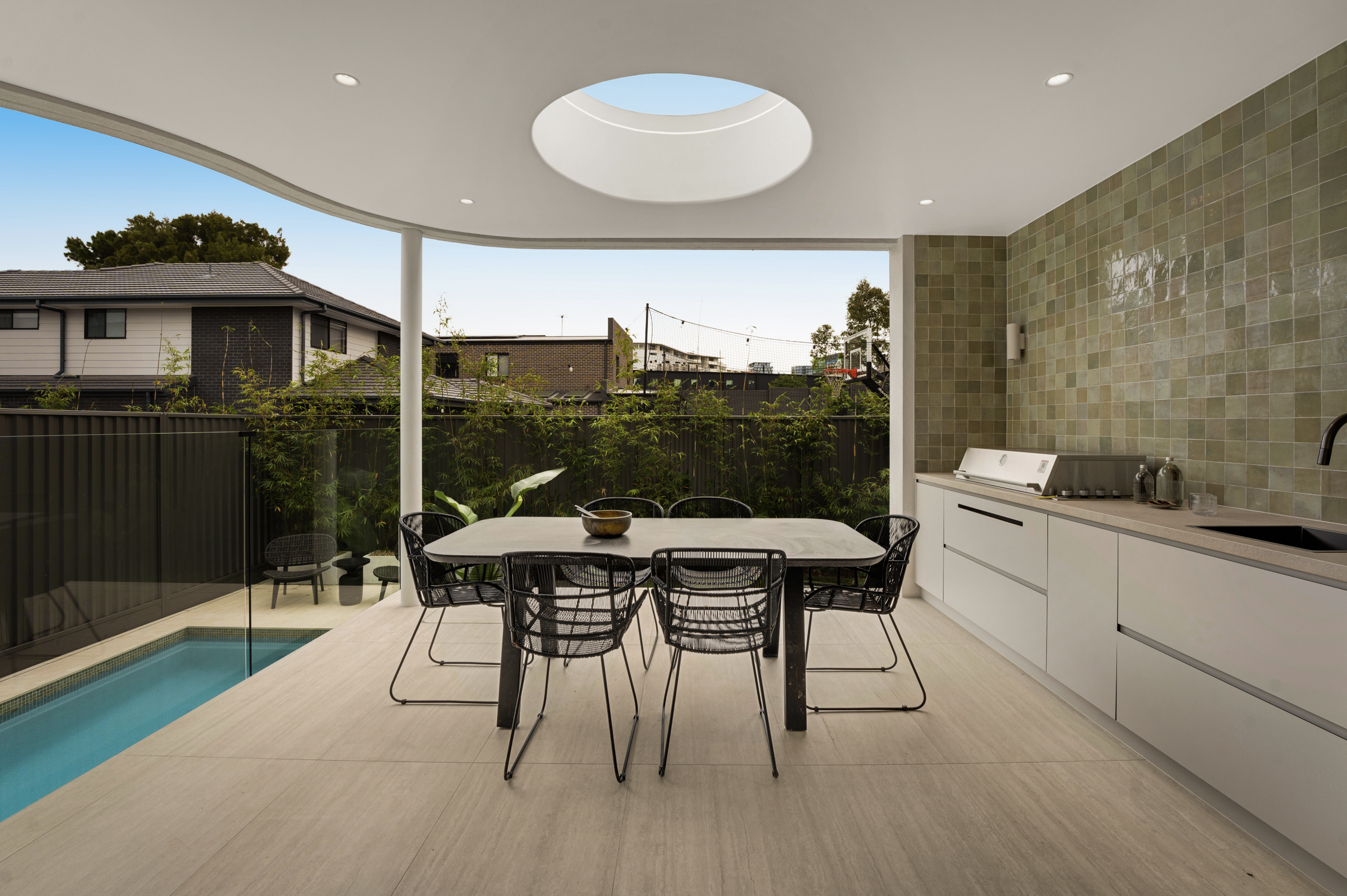 Modern outdoor patio with round dining table, black mesh chairs, built-in kitchen with green tiled wall, outdoor pool, privacy fencing, and neighboring houses with trees and basketball hoop in background.