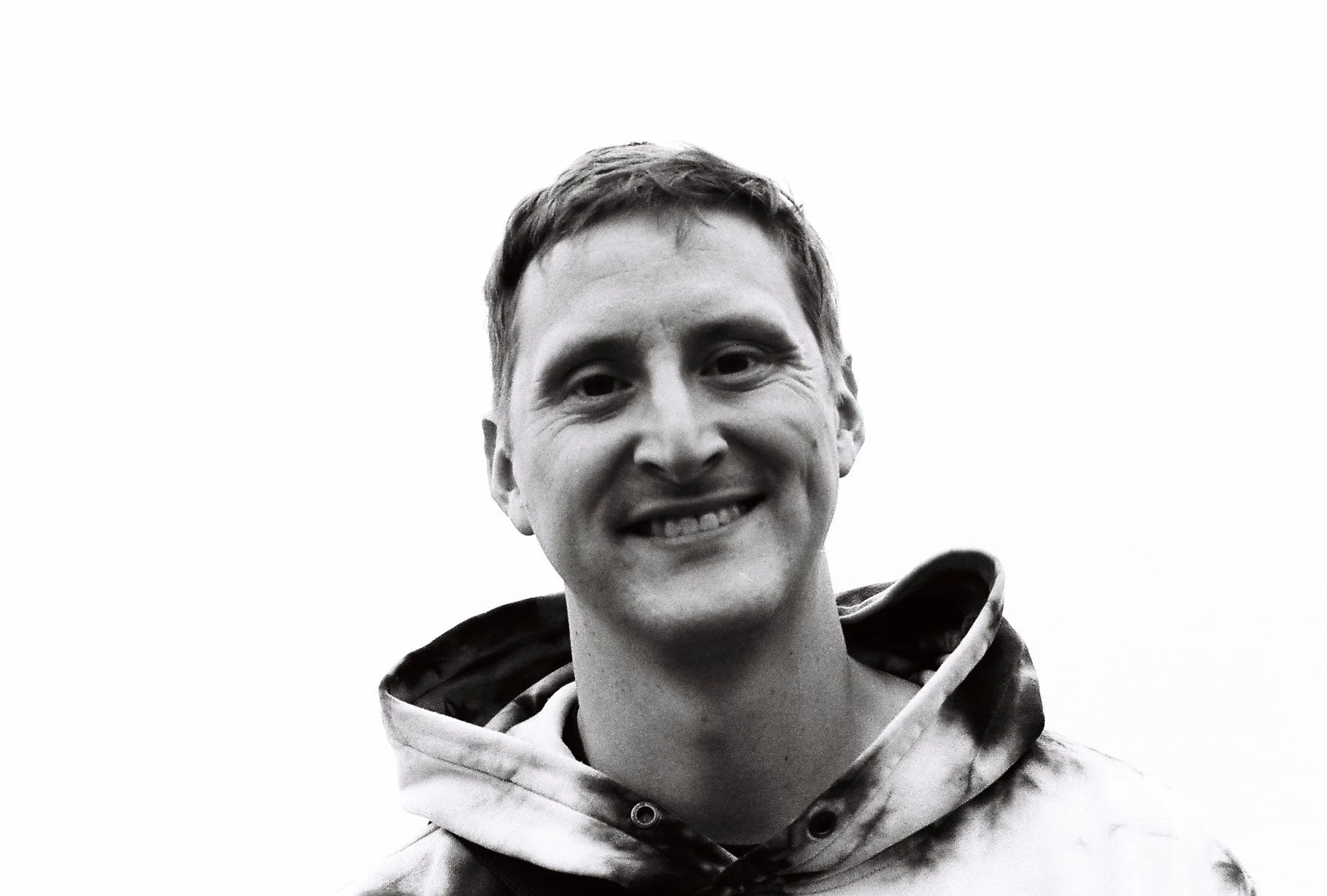 A black and white photo of a smiling young man wearing a hoodie, looking slightly to the side, with a light-colored sky background.