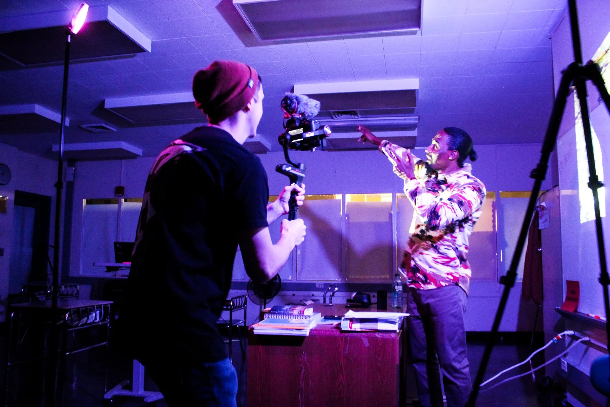 A man with dark hair, wearing a camoflauge  quarter zip , is filming or directing two other people who are talking to each other in a studio setting with colorful lighting and camera equipment.