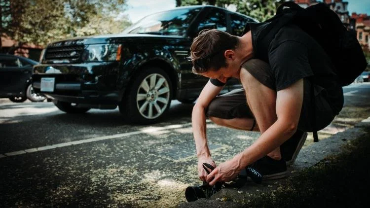 A young man crouching on the street, taking a low angle photo near a black SUV, on a city street during daytime.