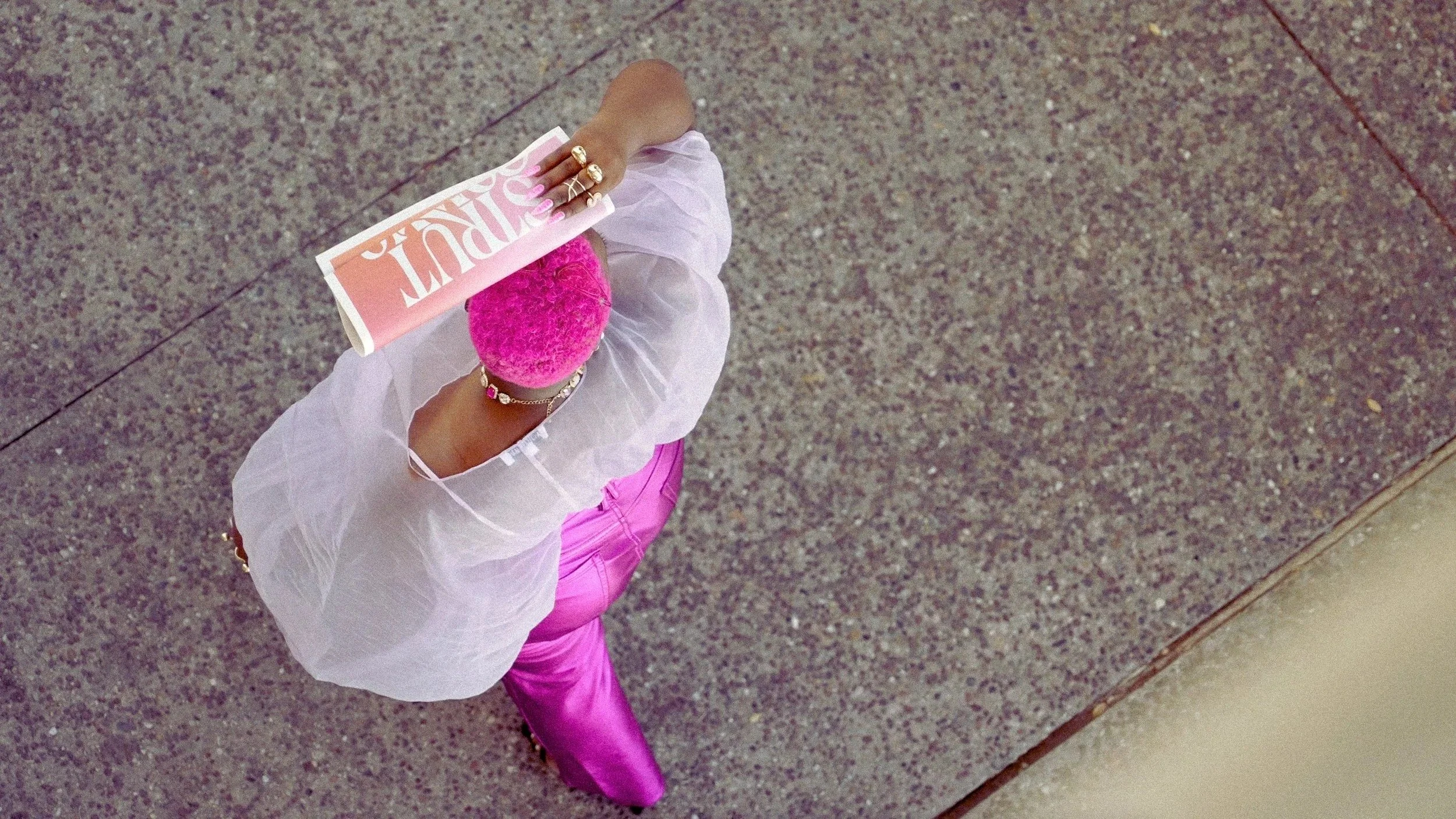 Overhead view of a person with pink hair, wearing a white top and bright pink pants, holding a magazine or book and walking on a sidewalk.