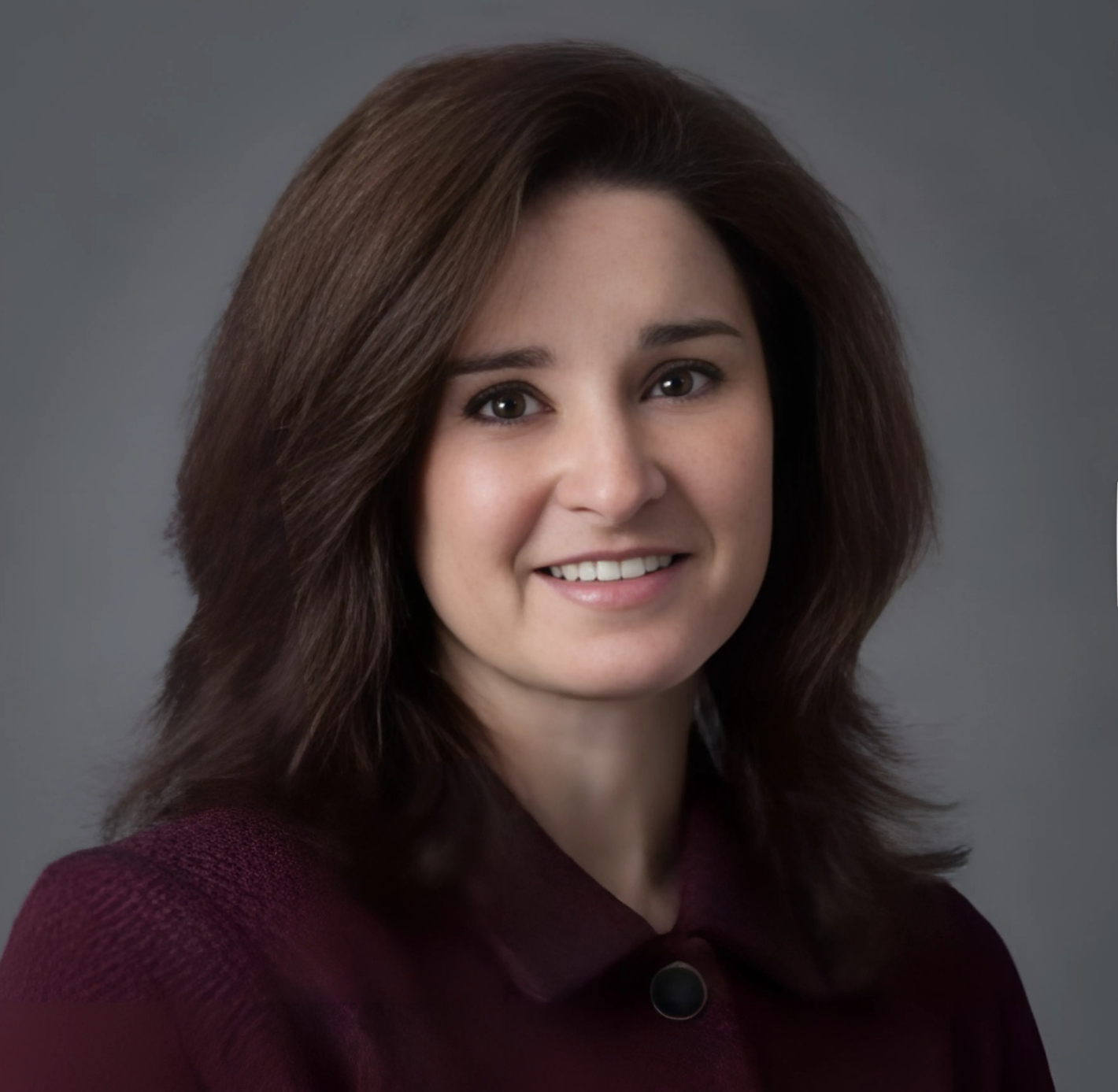 Photograph of a smiling woman with shoulder-length dark brown hair, wearing a dark maroon top, against a gray background.