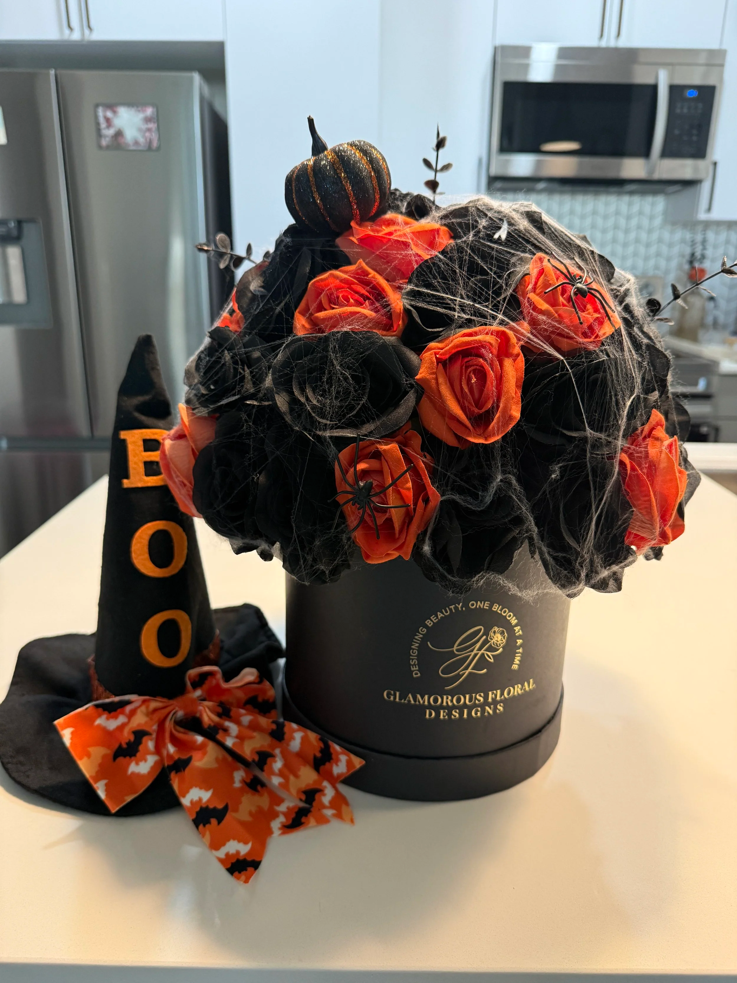 Black and orange faux roses arranged in a black hatbox with Halloween spiderwebs and decorative accents.