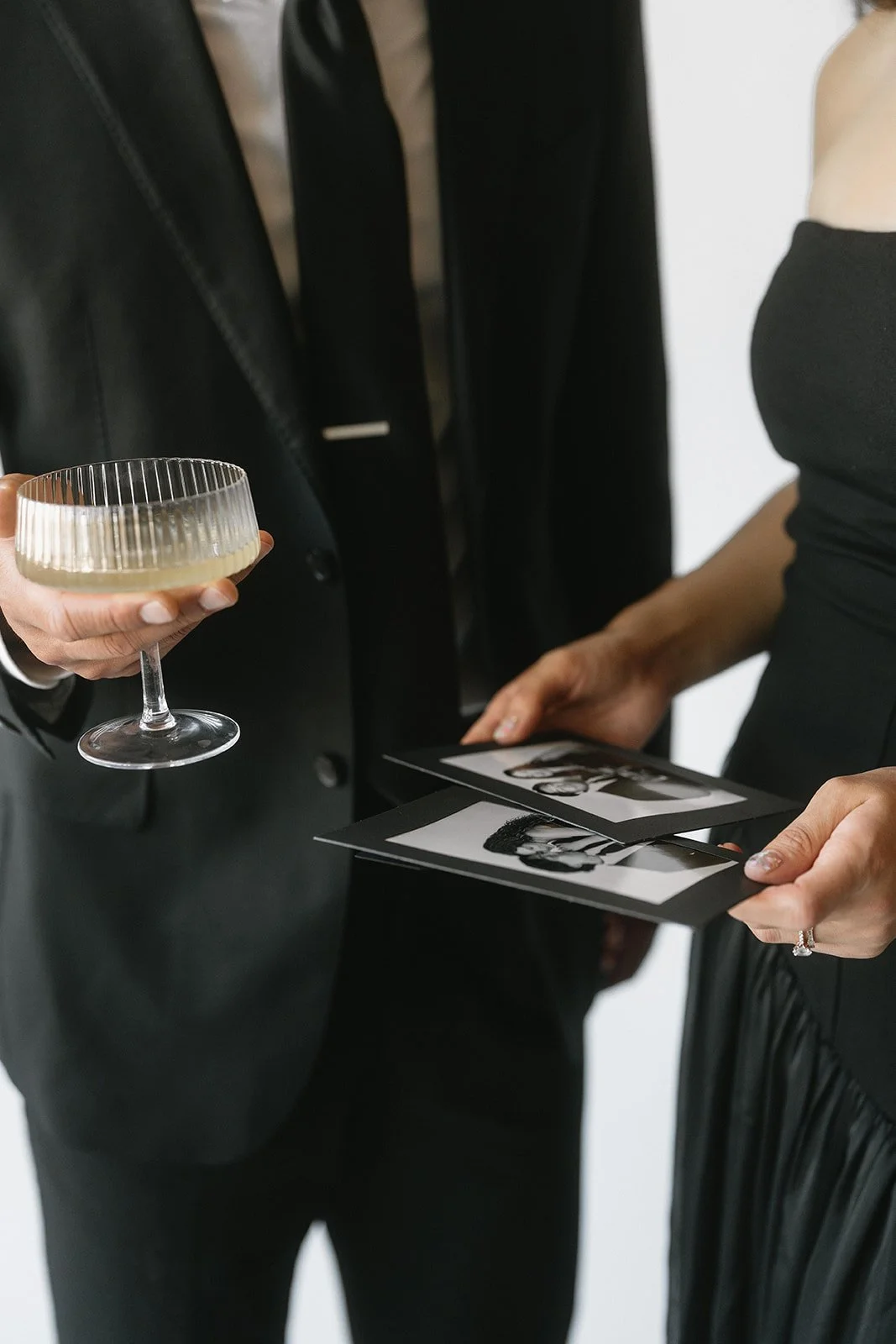 Two people, one in a black suit and the other in a black strapless dress, are engaging with black and white photographs, while the person in the suit holds a glass of champagne.