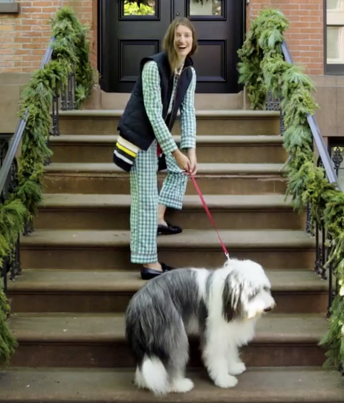Person in checkered pajamas and black vest walking a sheepdog on stone steps with green garlands on the railing.