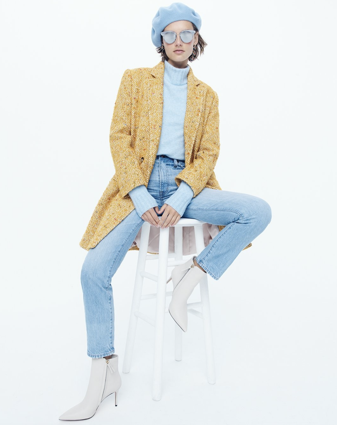 Fashionable woman wearing a yellow coat, blue beret, turtleneck sweater, jeans, and white boots, sitting on a stool against a plain background.