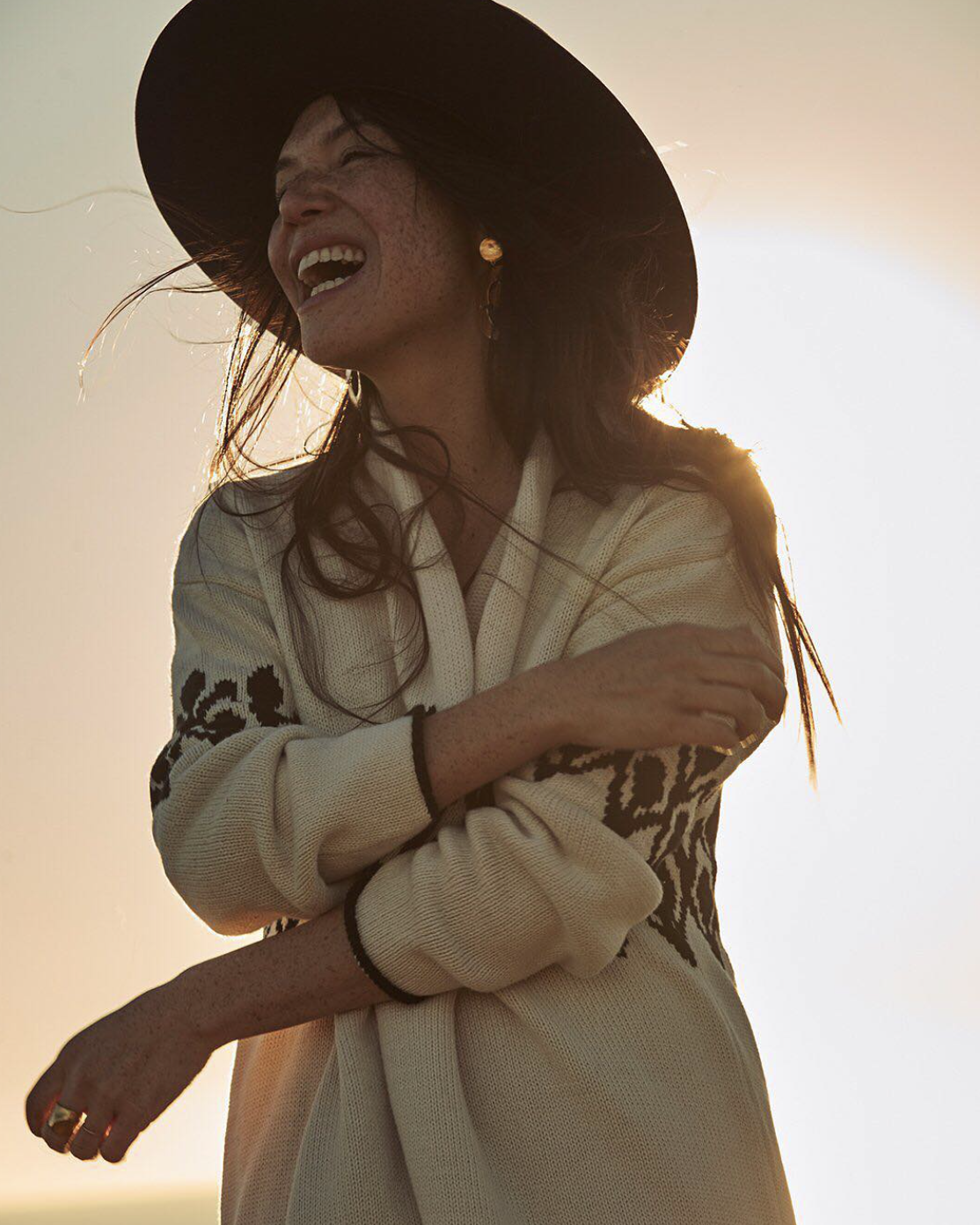 Smiling woman in a wide-brimmed hat and patterned sweater, backlit by the sun.