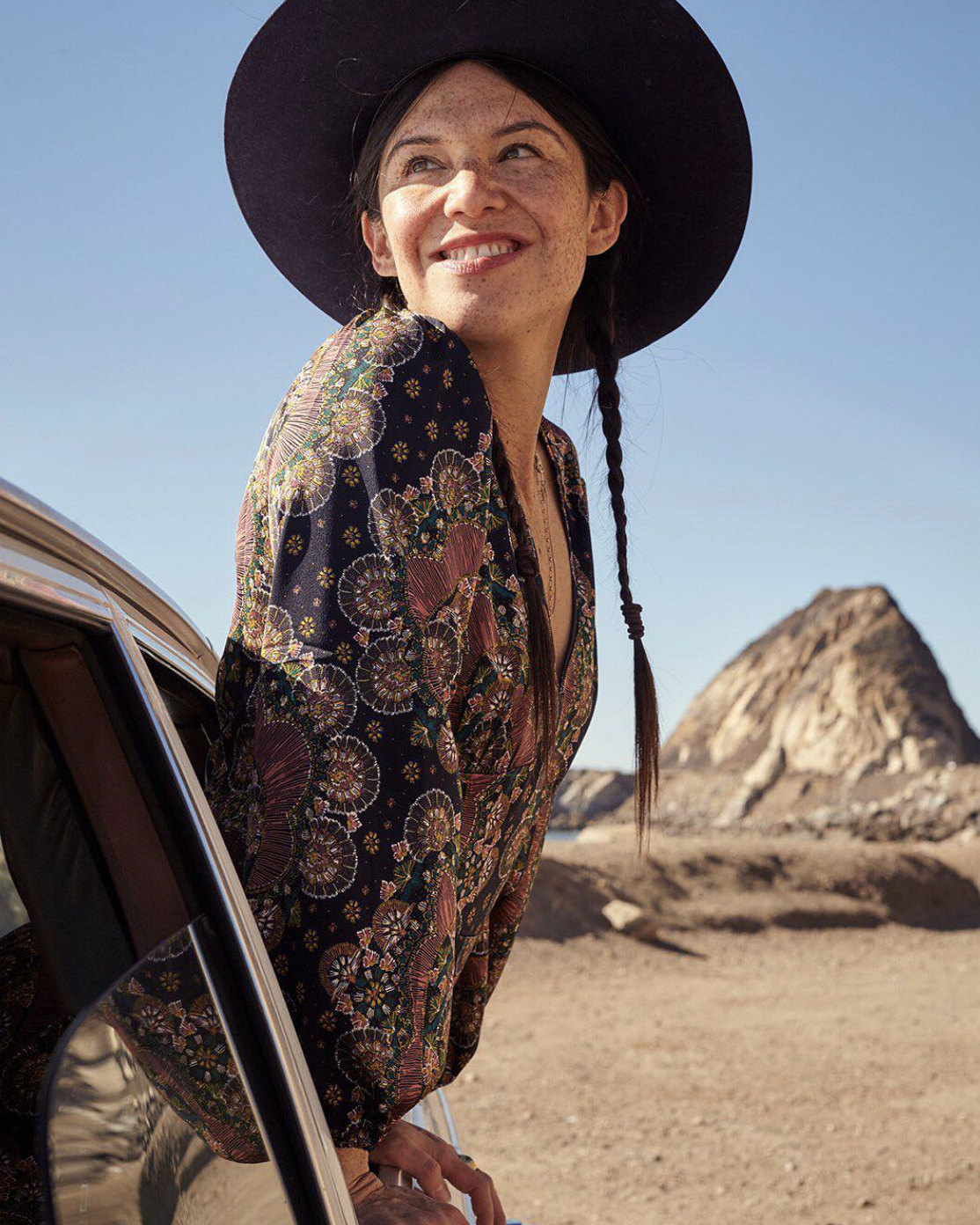 Woman in patterned outfit and wide-brimmed hat leaning out of car window in a desert landscape.