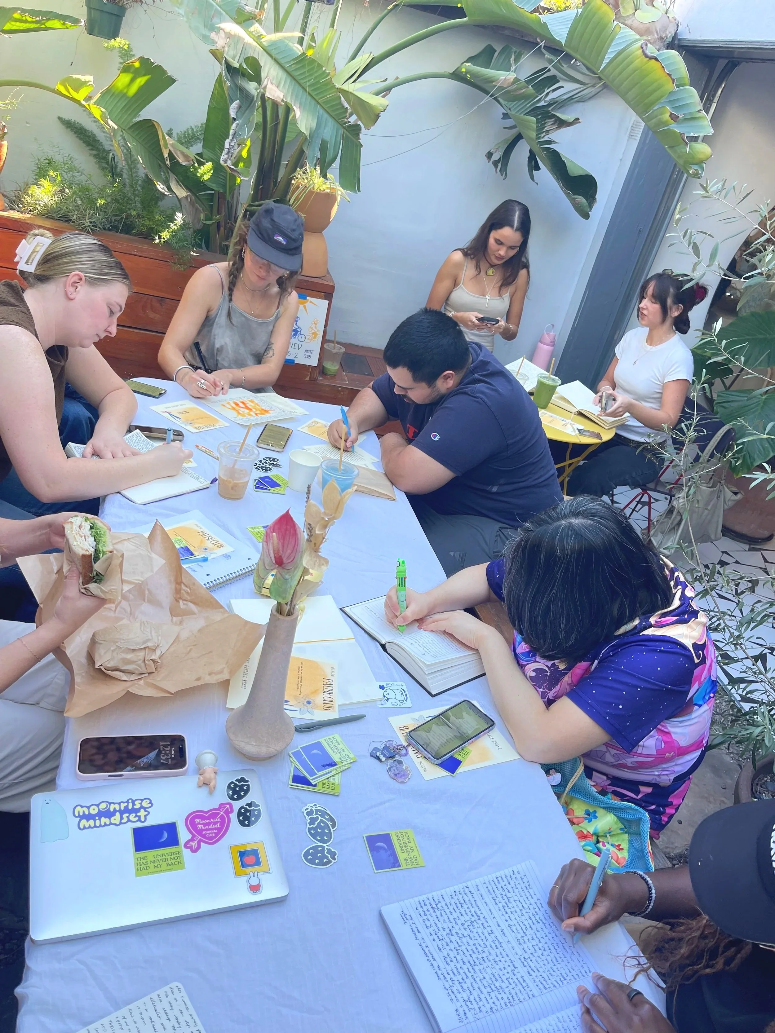People gathered around a table outdoors, engaged in writing, reading, and discussing, with notebooks, books, and drinks on the table, surrounded by green plants.