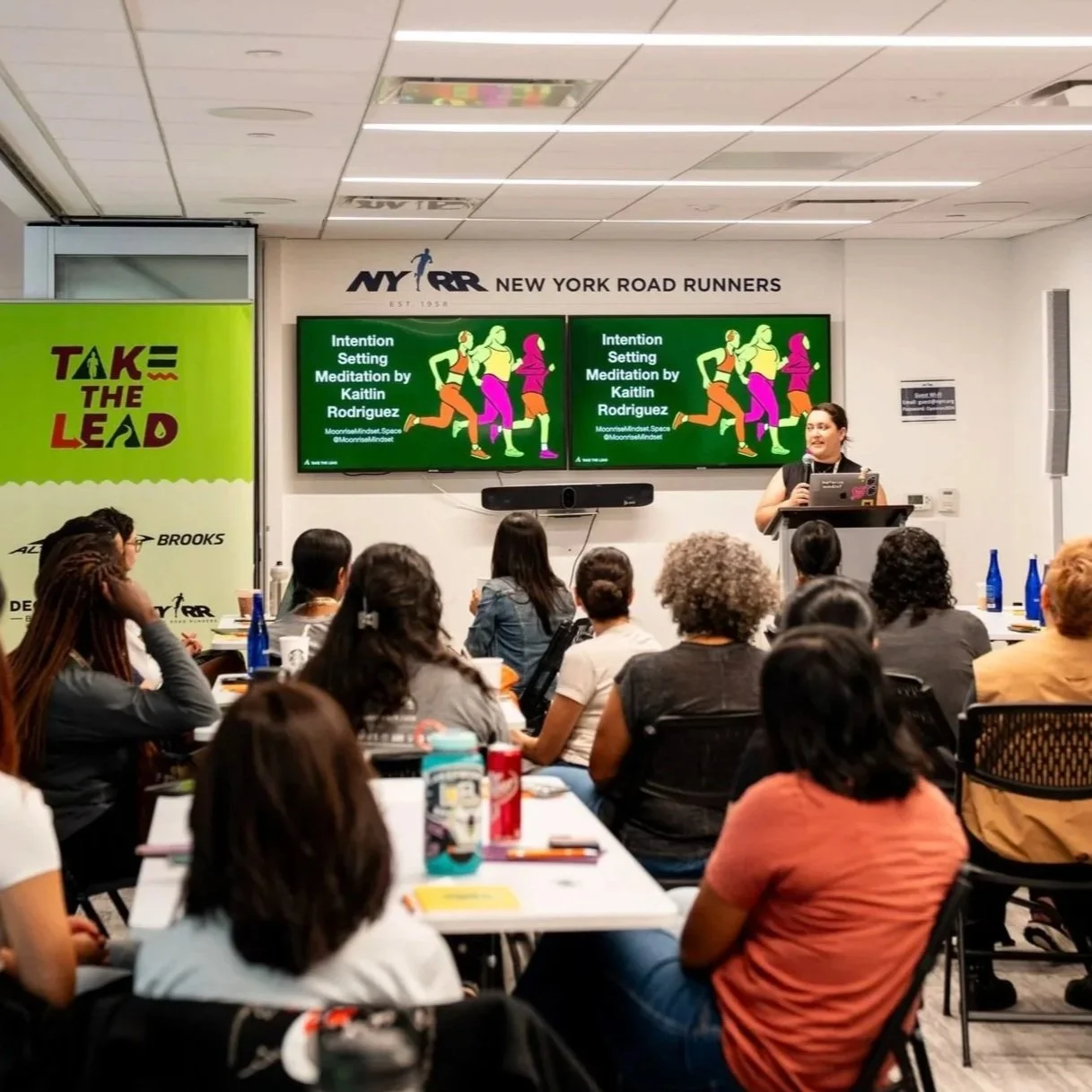 Woman giving a presentation at a conference with audience sitting at tables, two digital screens displaying a presentation titled 'Intention Setting Meditation by Kaitlin Rodriguez,' and a banner with the words 'Take the Lead' on the left side.