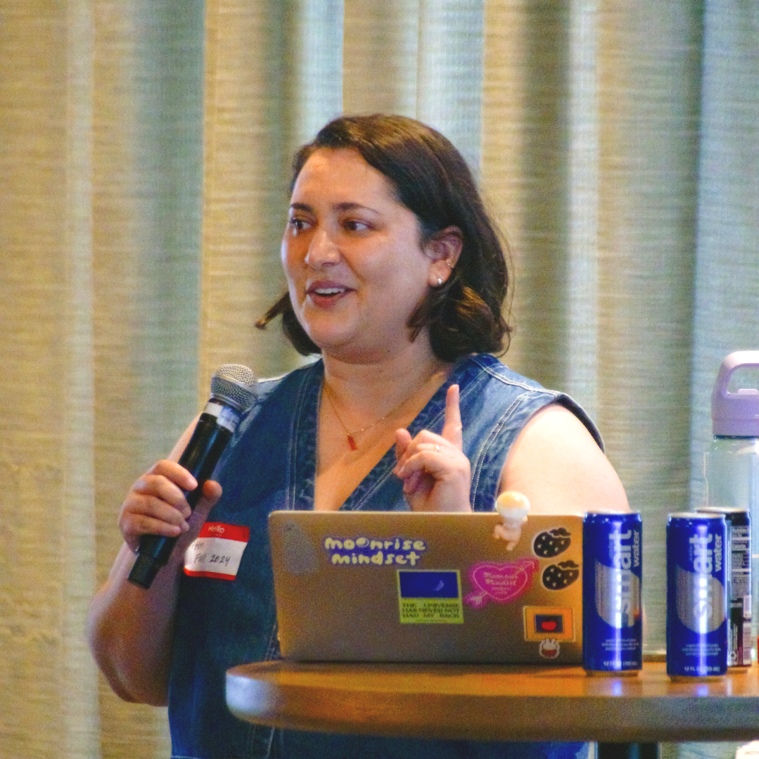 A woman with dark hair, wearing a sleeveless denim vest, holding a microphone and speaking, standing behind a round table with a laptop covered in colorful stickers, and two cans of Red Bull energy drinks nearby.