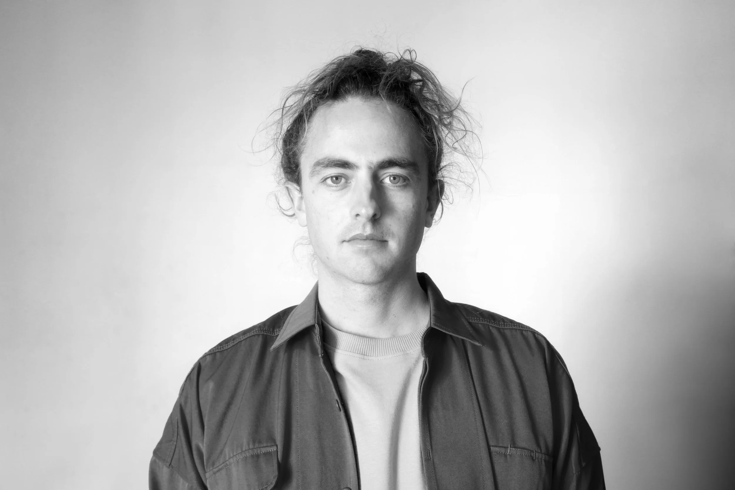 Black and white portrait of a young man with curly hair, wearing a jacket over a t-shirt, looking directly at the camera against a plain background.