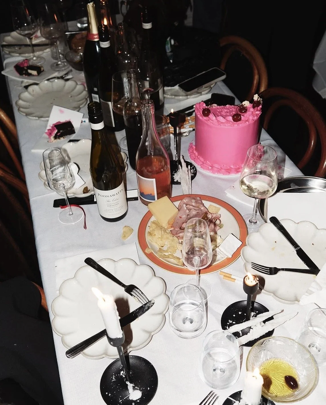 A dinner table with plates, wine glasses, bottles of wine, a pink birthday cake with cherries, candles, a cheese and meat platter, and assorted drinks and utensils.