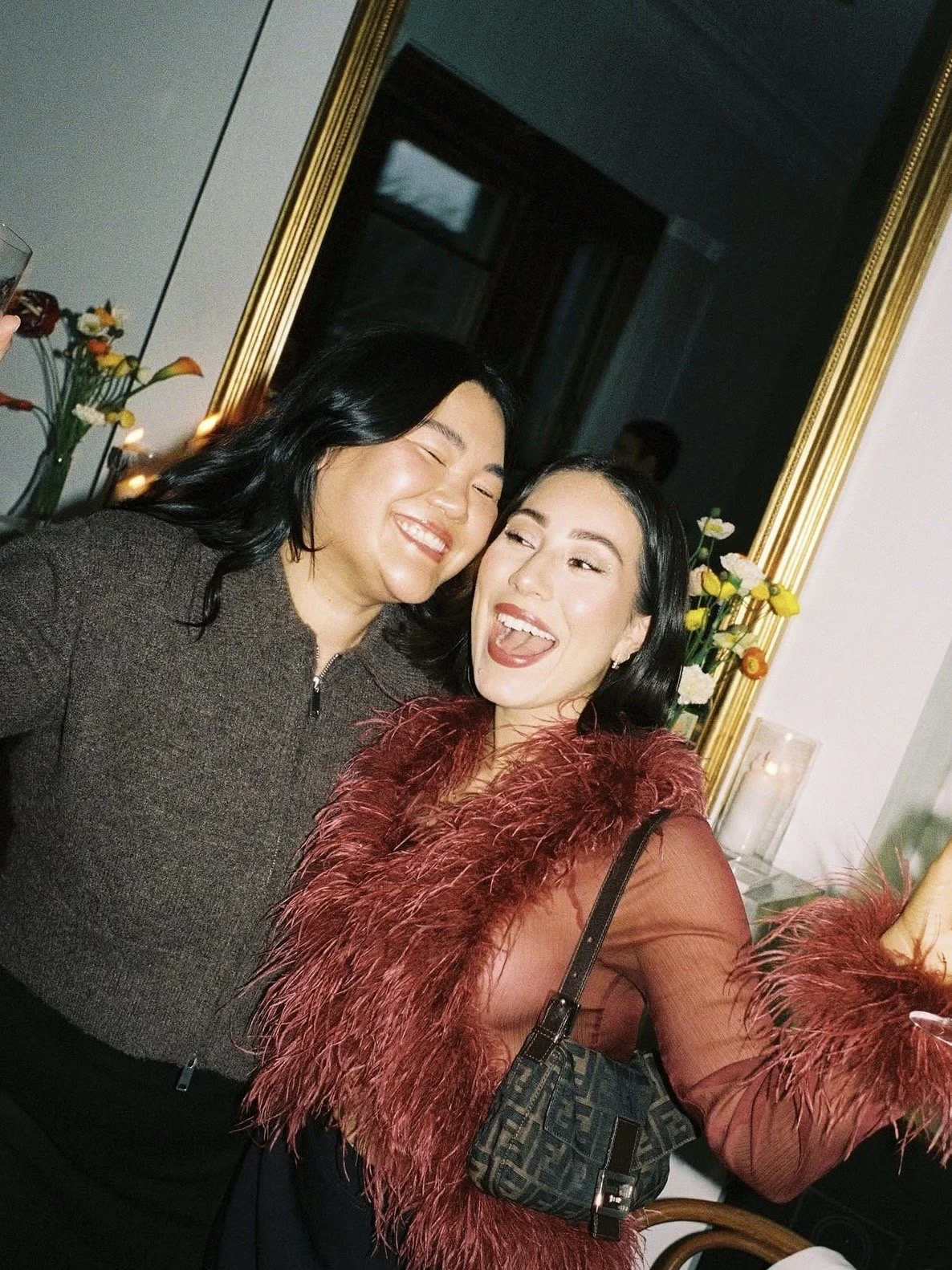 Two women smiling and taking a selfie at a party or gathering, with a mirror and flowers in the background.