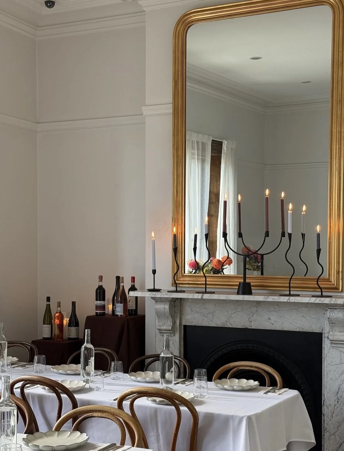 Elegant dining room with a white tablecloth, glass bottles, and place settings. A large mirror with a gold frame above a marble fireplace reflects a window with white curtains. Candleholders with lit candles are on the fireplace.