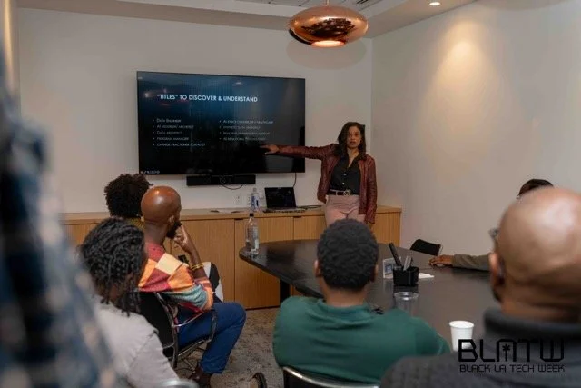 A woman giving a presentation to a group of people in a conference room, with a screen displaying a slide titled 'Titles to Discover & Understand'.