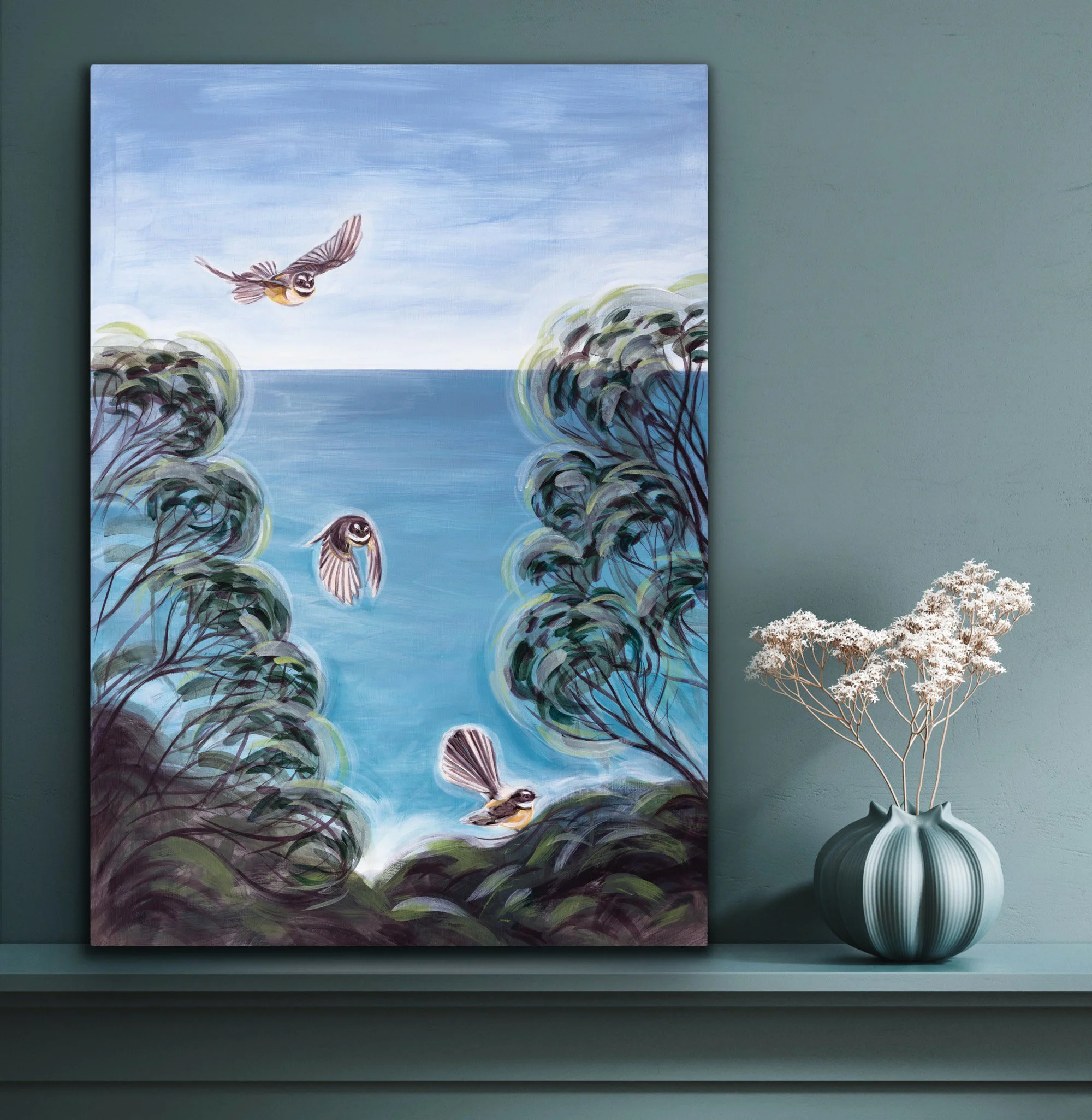 A painting of four birds flying over a blue water scene framed by trees, hanging on a green wall next to a vase with white flowers.