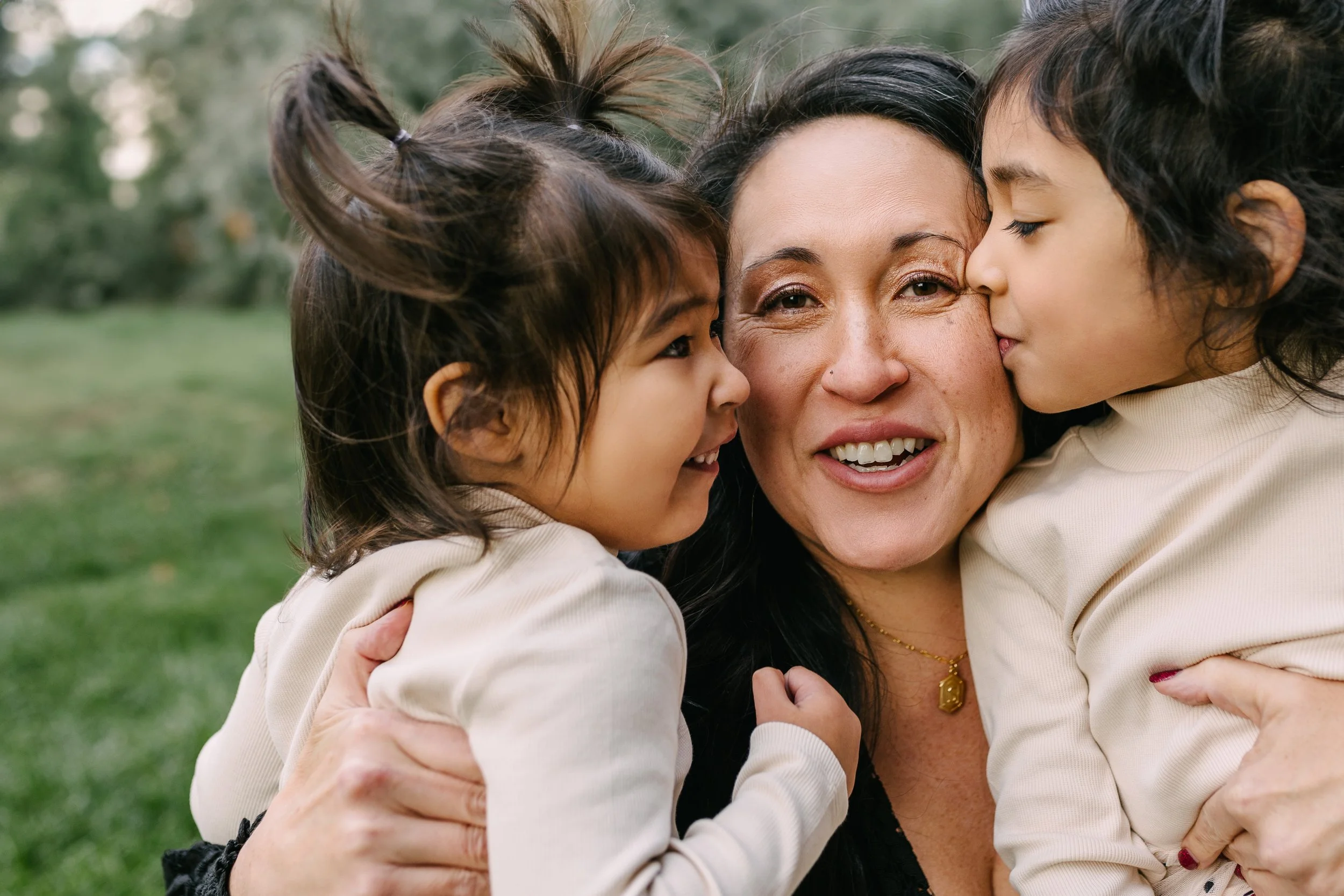 Mom smiles at the camera while her two young daughters kiss her cheeks during a candid fall family photo session in Overland Park, KS.