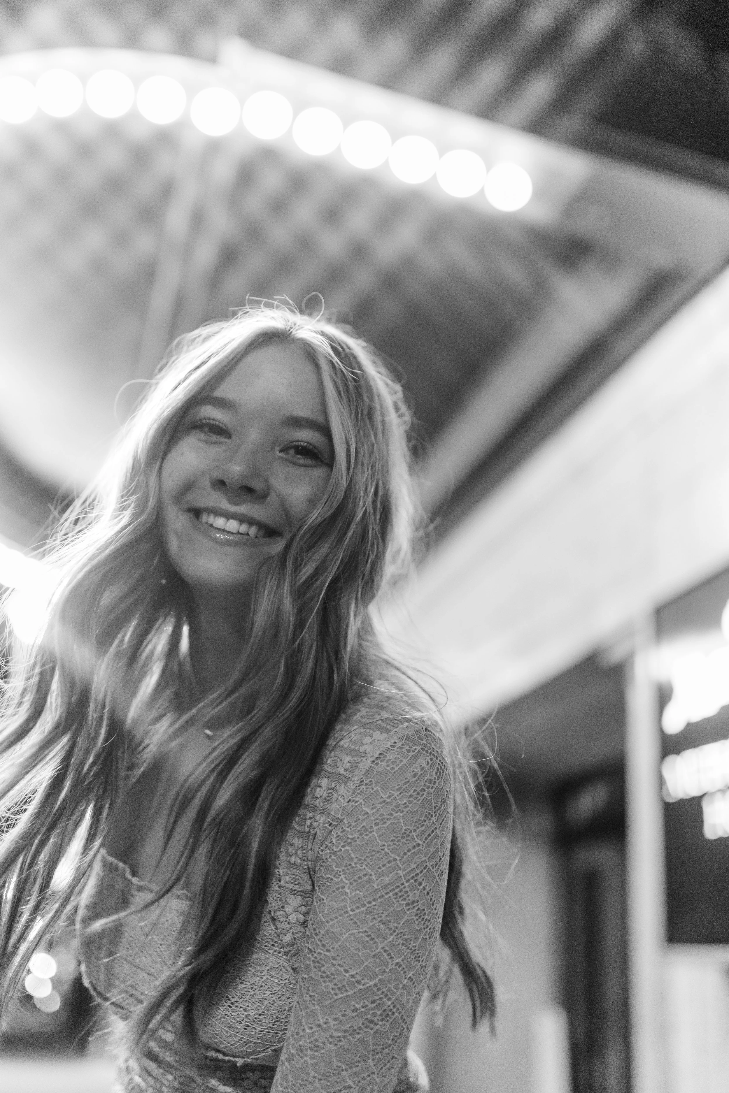 A high school senior smiles at the camera under vintage marquee lights during a nighttime editorial senior portrait session in downtown Kansas City, MO.