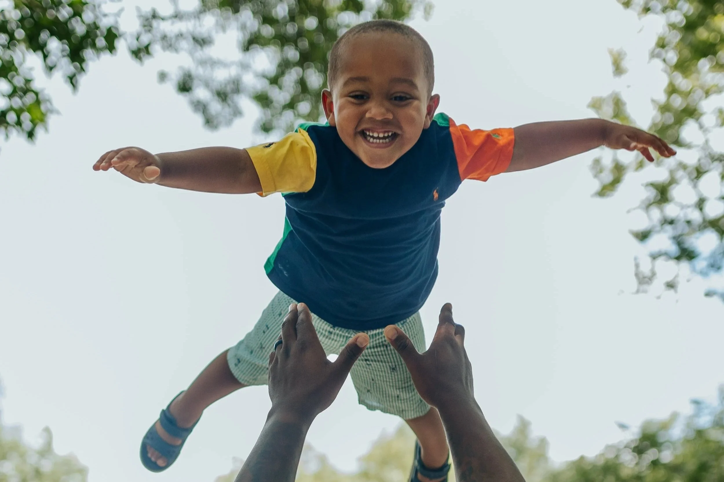 Dad throws young boy into the air while he smiles at the camera with arms outstretched during candid family photo session in Leawood, KS.