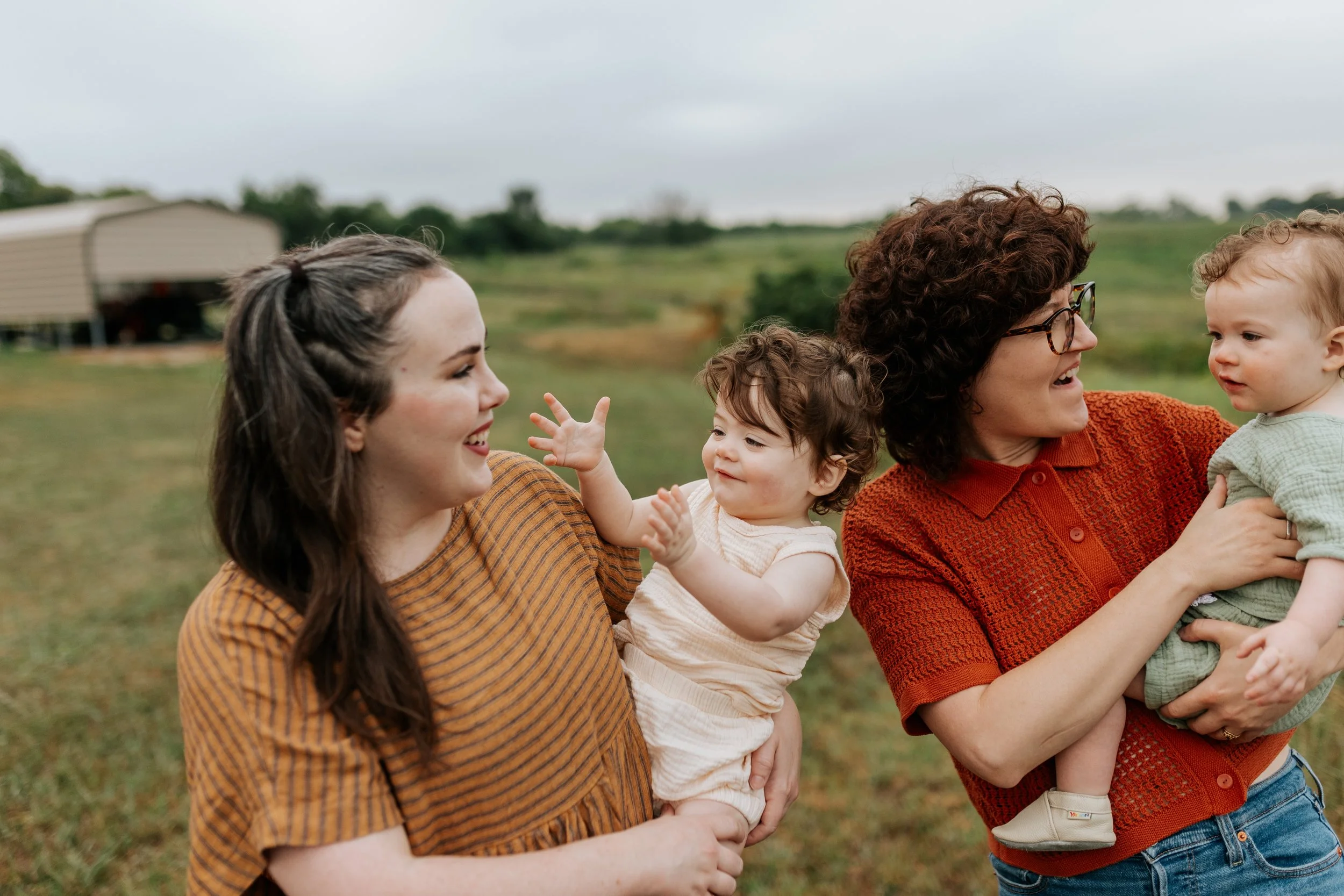 Two moms hold their twin toddlers as they walk through a grass field during an early morning candid family photo session at the prairie center in Olathe, KS.