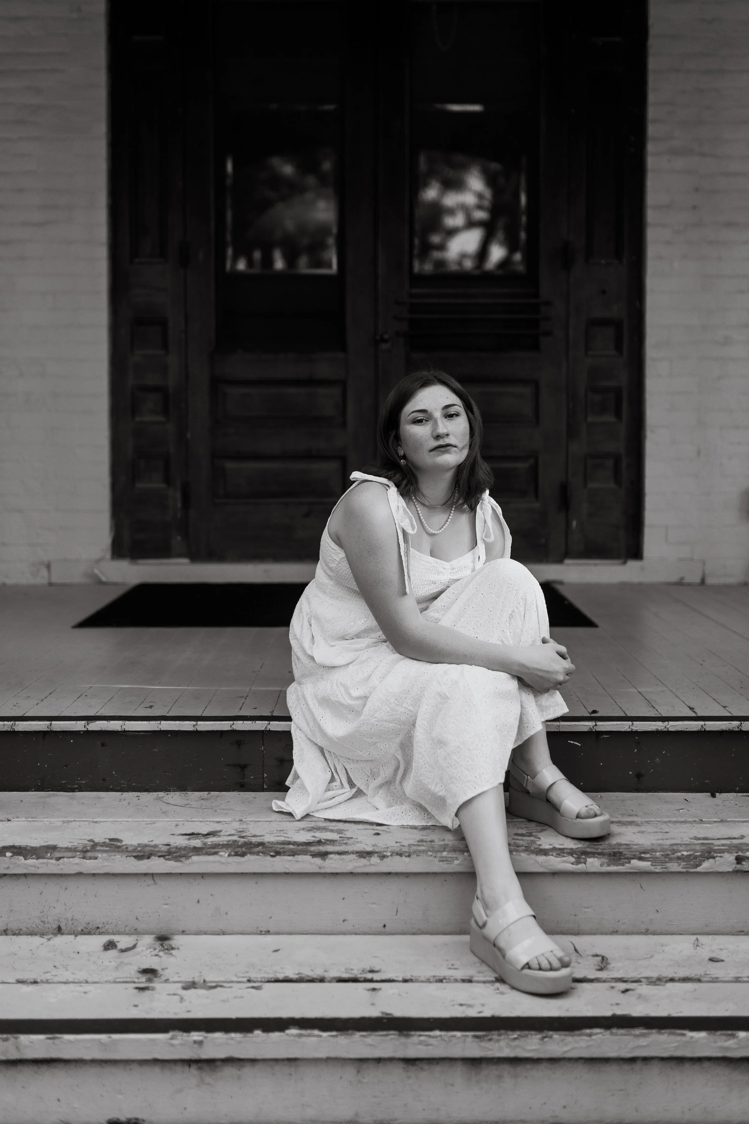 A high school senior looks at the camera while sitting on the porch of the Victorian Bingham-Waggoner Estate while hugging her knee during an editorial storytelling senior photo session.