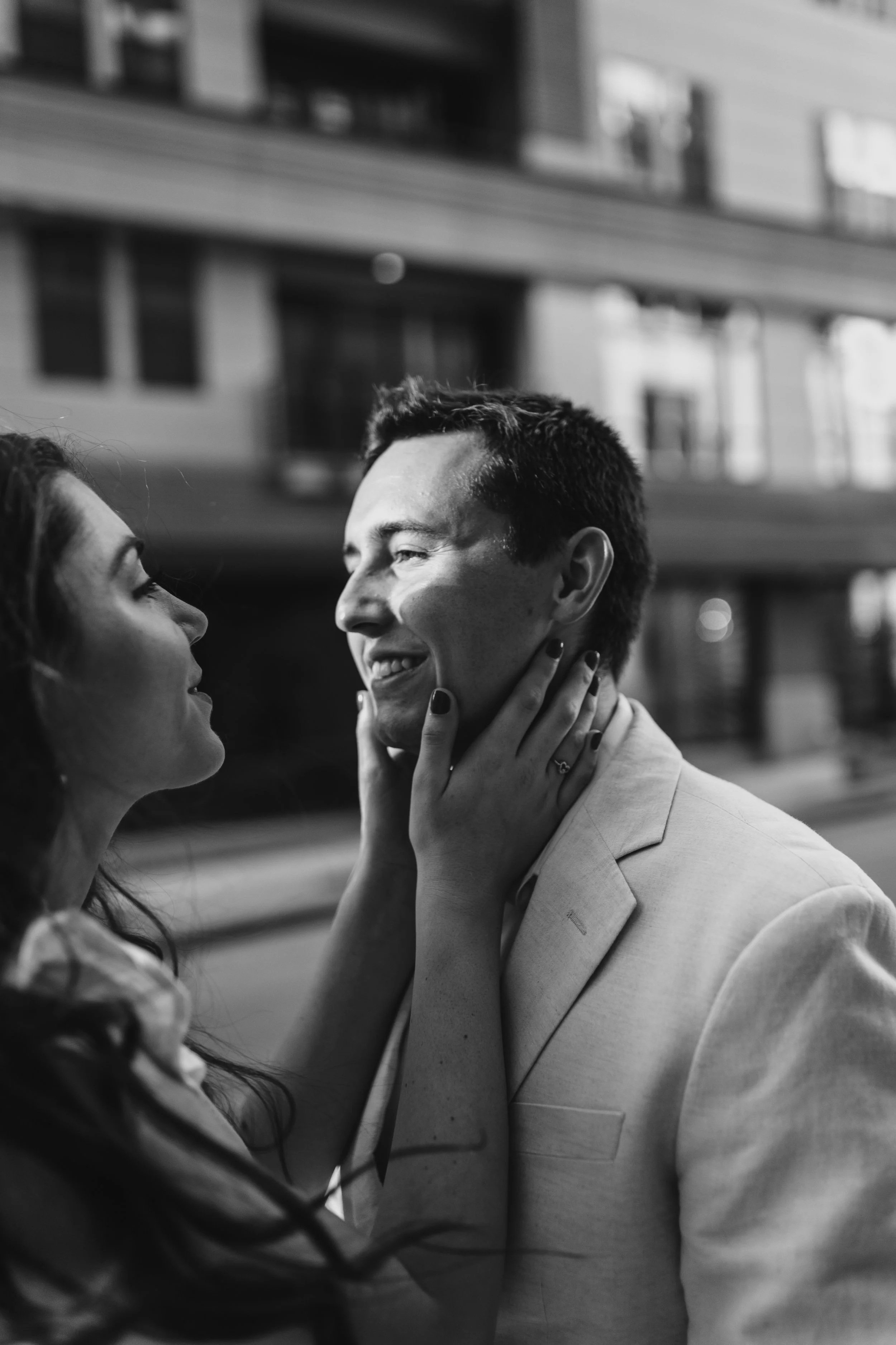 An engaged couple smile at each other while the bride holds the groom's face during a candid summer engagement photoshoot on the street in downtown Kansas City, MO.