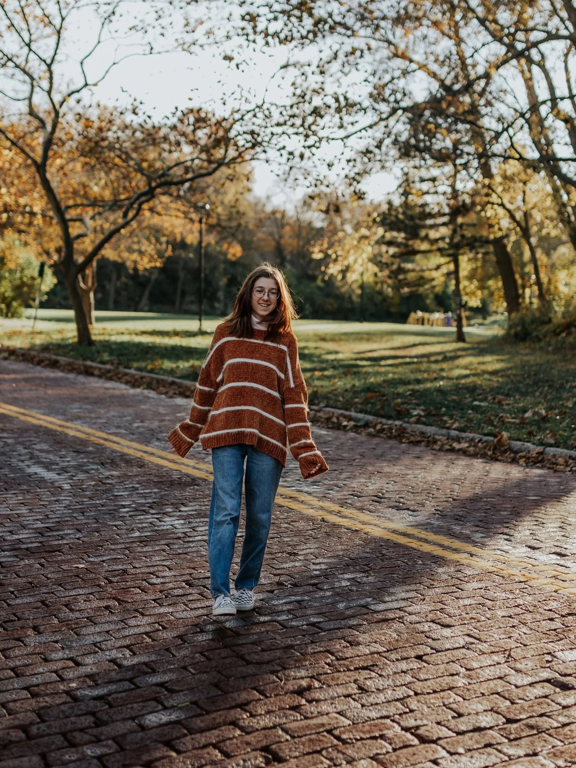 A high school senior walks across a red brick road at golden hour during a cozy fall senior photoshoot at Roanoke Park in Kansas City.