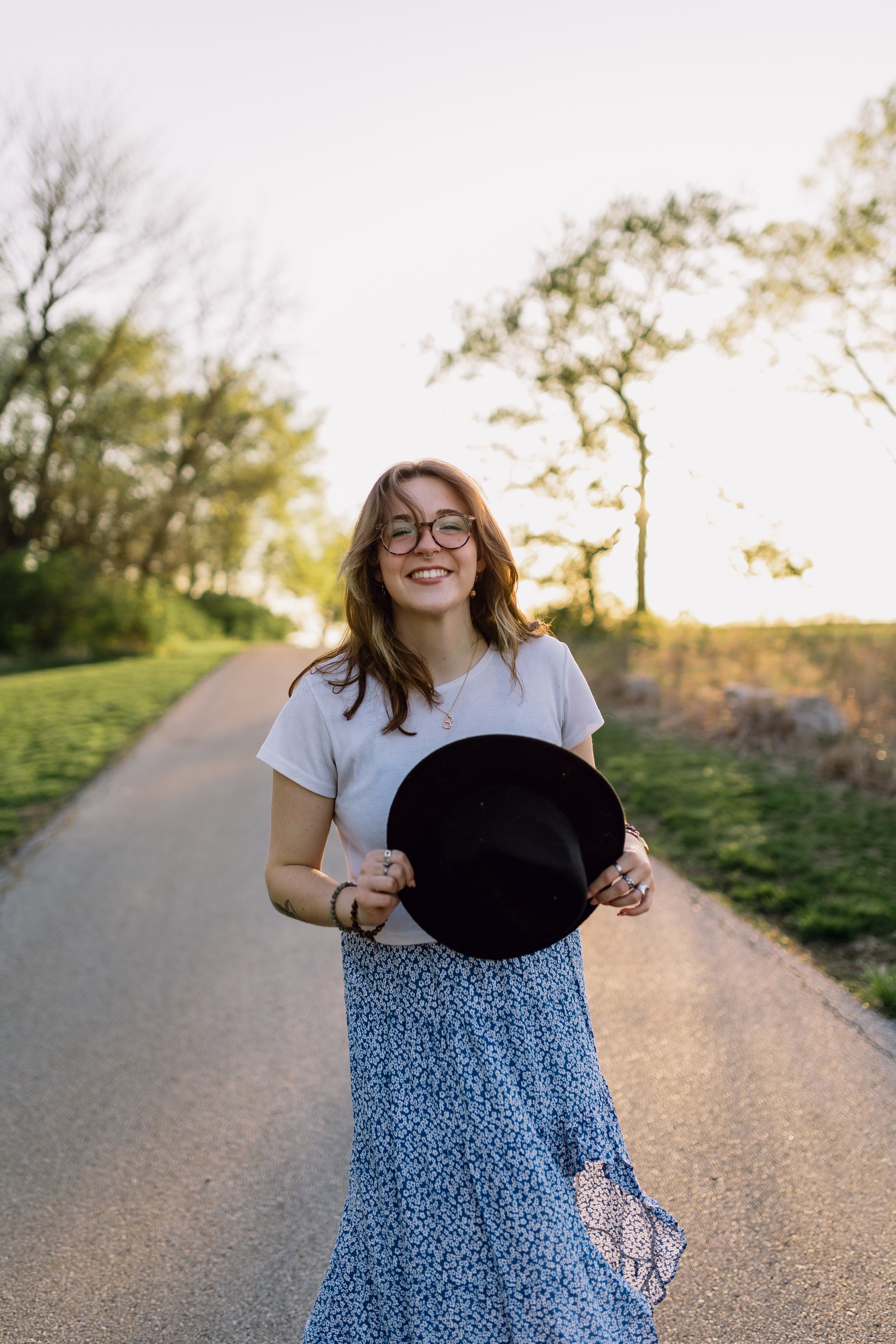 A high school senior in a flowy boho outfit smiles at the camera while twirling on a paved trail during a golden hour senior photoshoot at Jerry Smith Park in south Kansas City, MO.