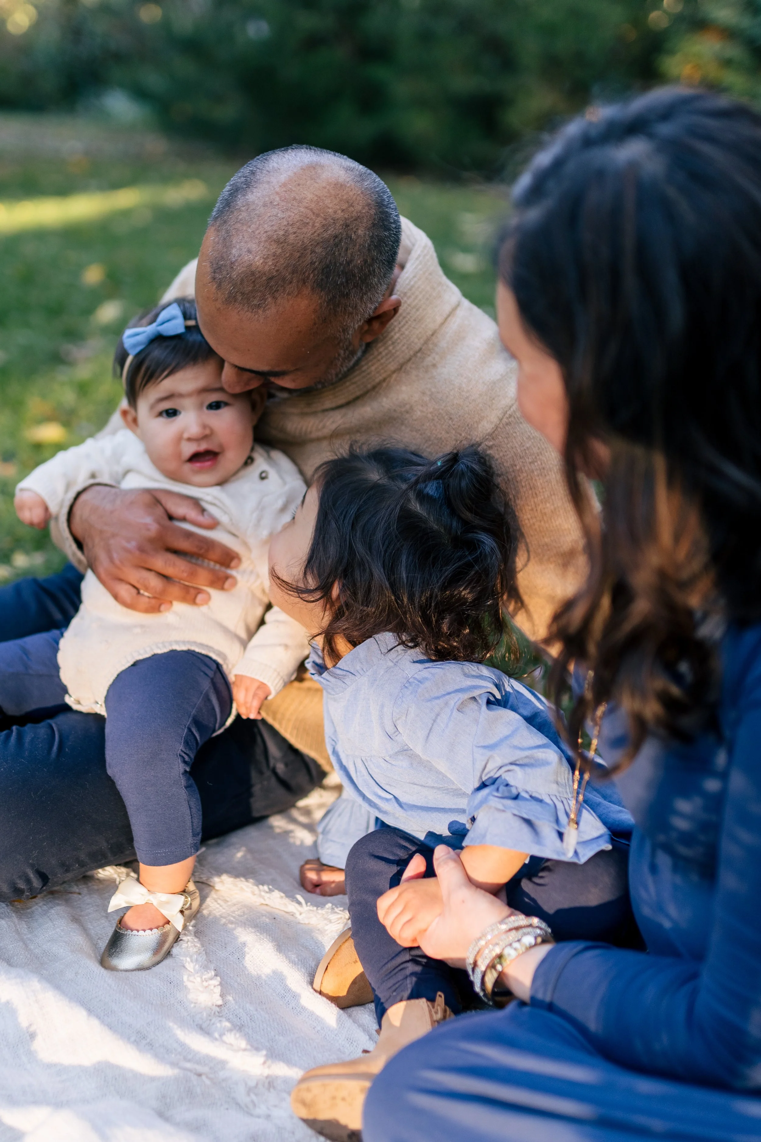 A mom, dad, and toddler smile at the baby of the family while the baby smiles at the camera during a candid backyard at home family photoshoot in Leawood, KS.