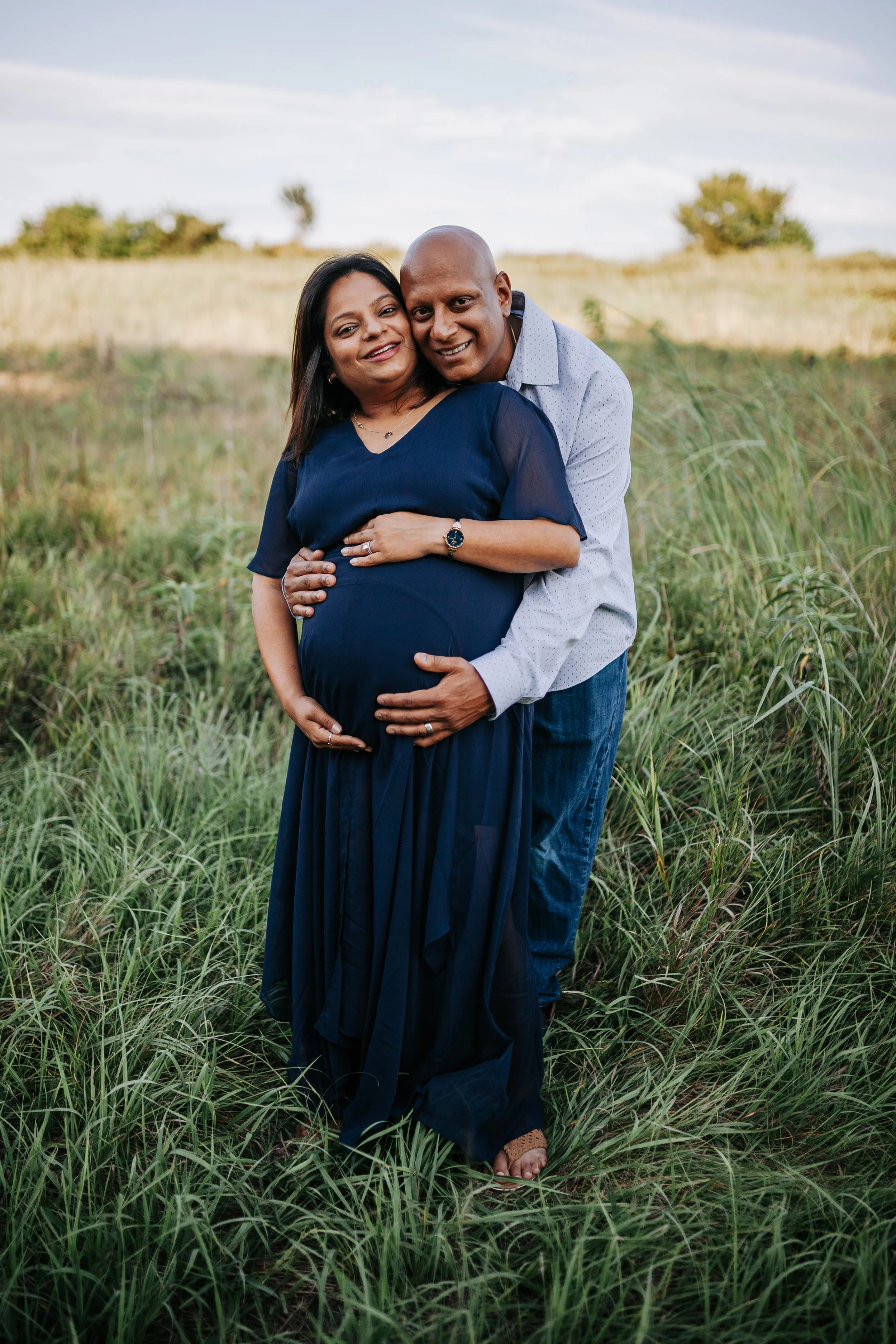A pregnant woman stands in front of her husband as they both cradle her bump during a summer maternity photo session at the prairie center park in Olathe, KS.