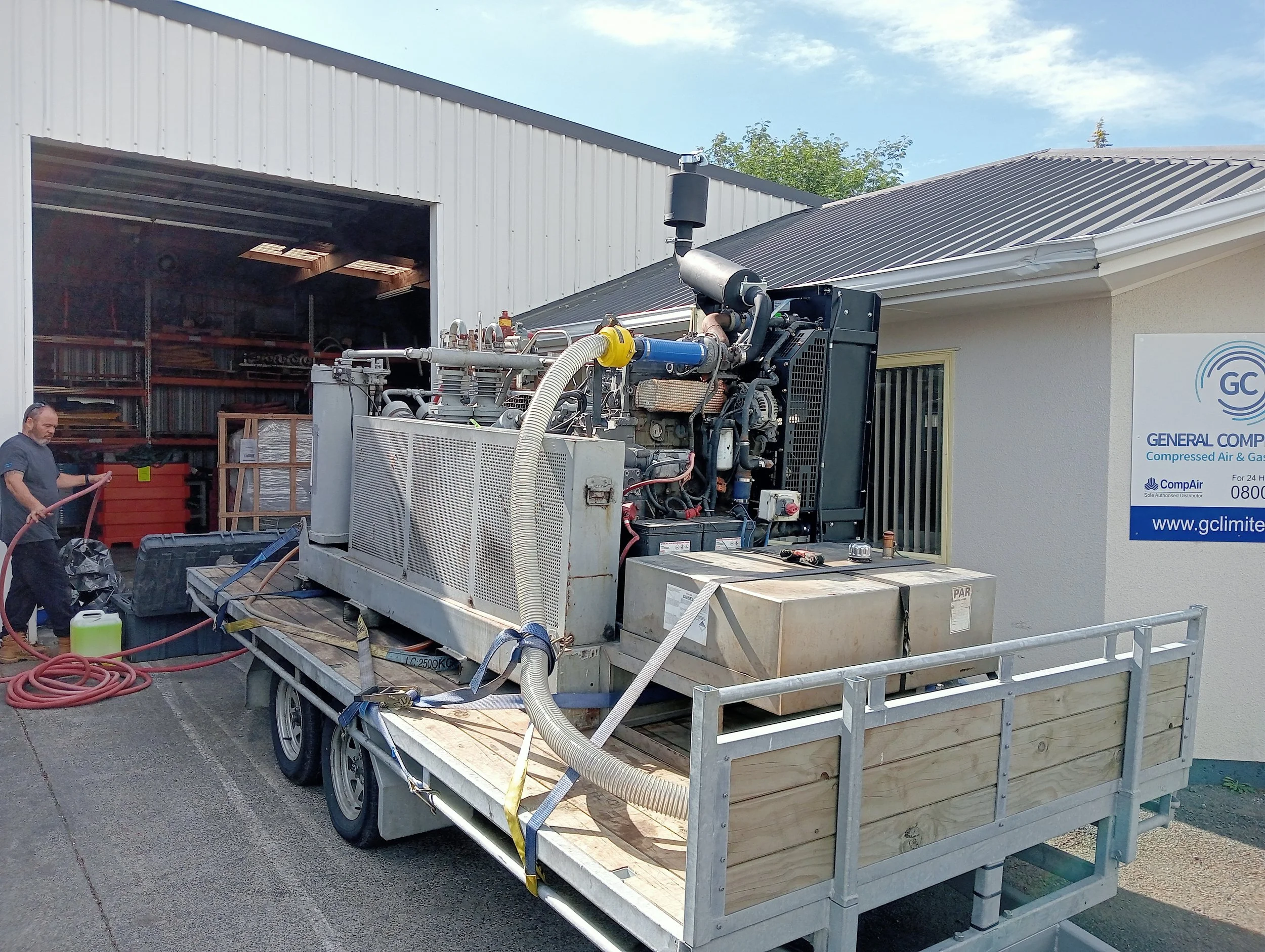 A man working with a hose next to a large industrial machine on a trailer outside a warehouse building.
