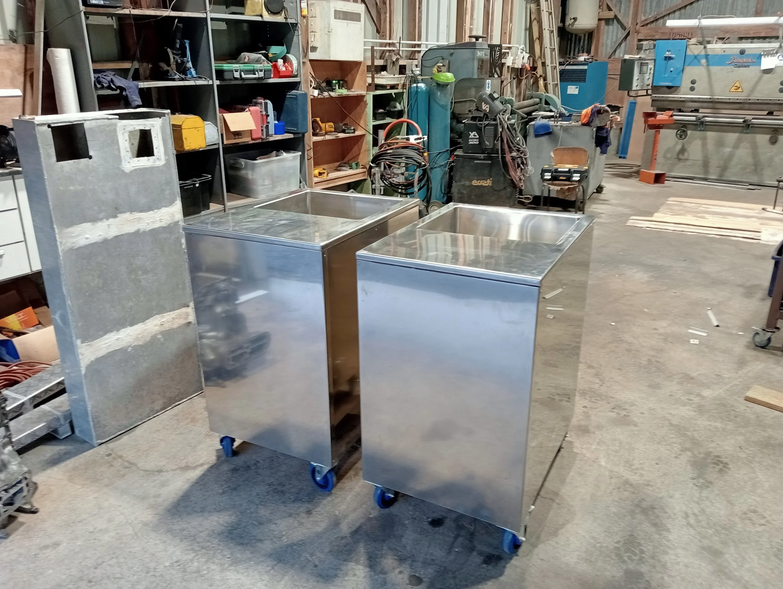Two stainless steel mobile storage carts with blue wheels in a workshop or industrial setting.