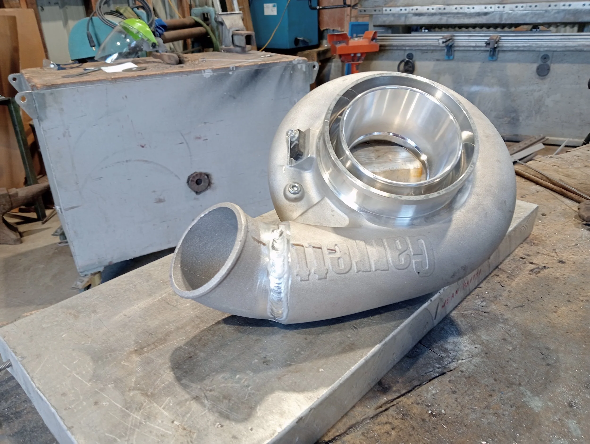 A turbocharger with a shiny metal compressor wheel is placed on a workbench in a workshop, with tools and equipment in the background.