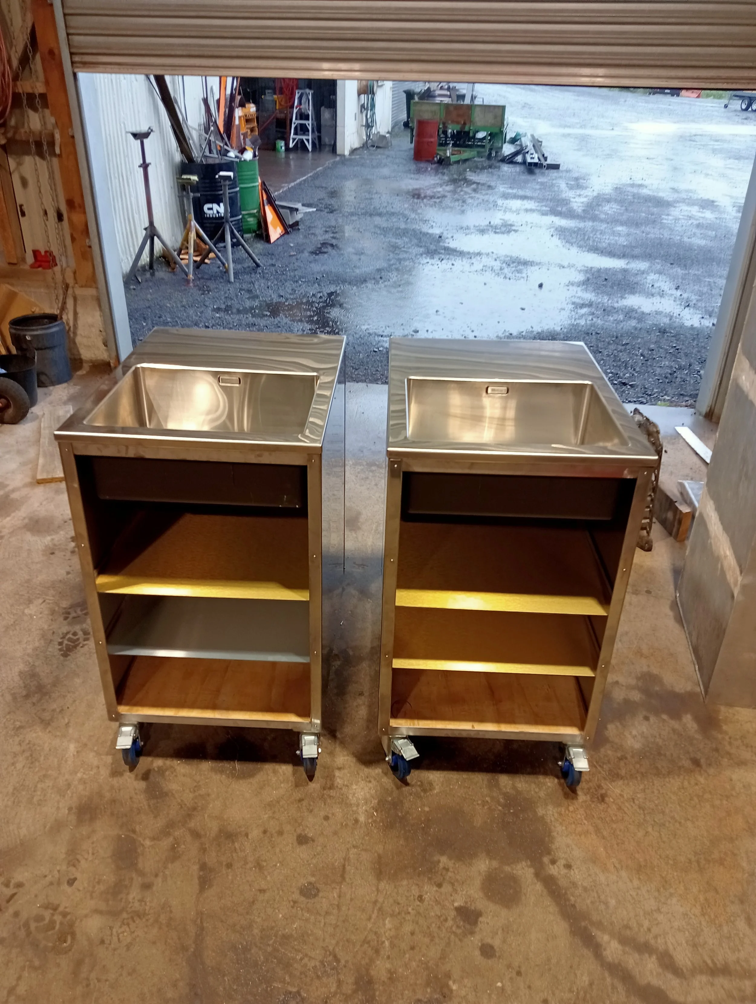 Two stainless steel utility carts with sinks, each with two shelves underneath, on caster wheels in a workshop or garage setting with an open garage door revealing a wet outdoor area with tools and equipment.