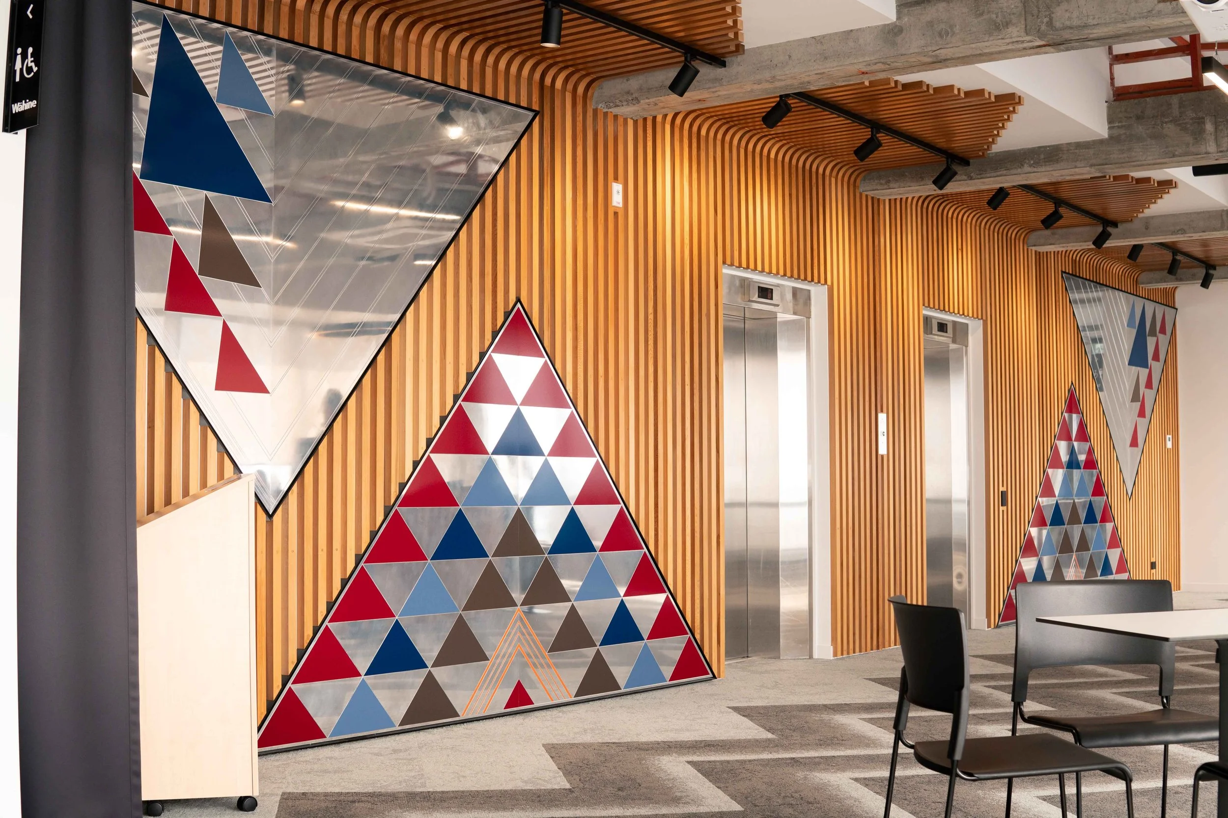 Interior of a modern building with wooden slat wall decorations and geometric artwork, including triangles, near two elevator doors.