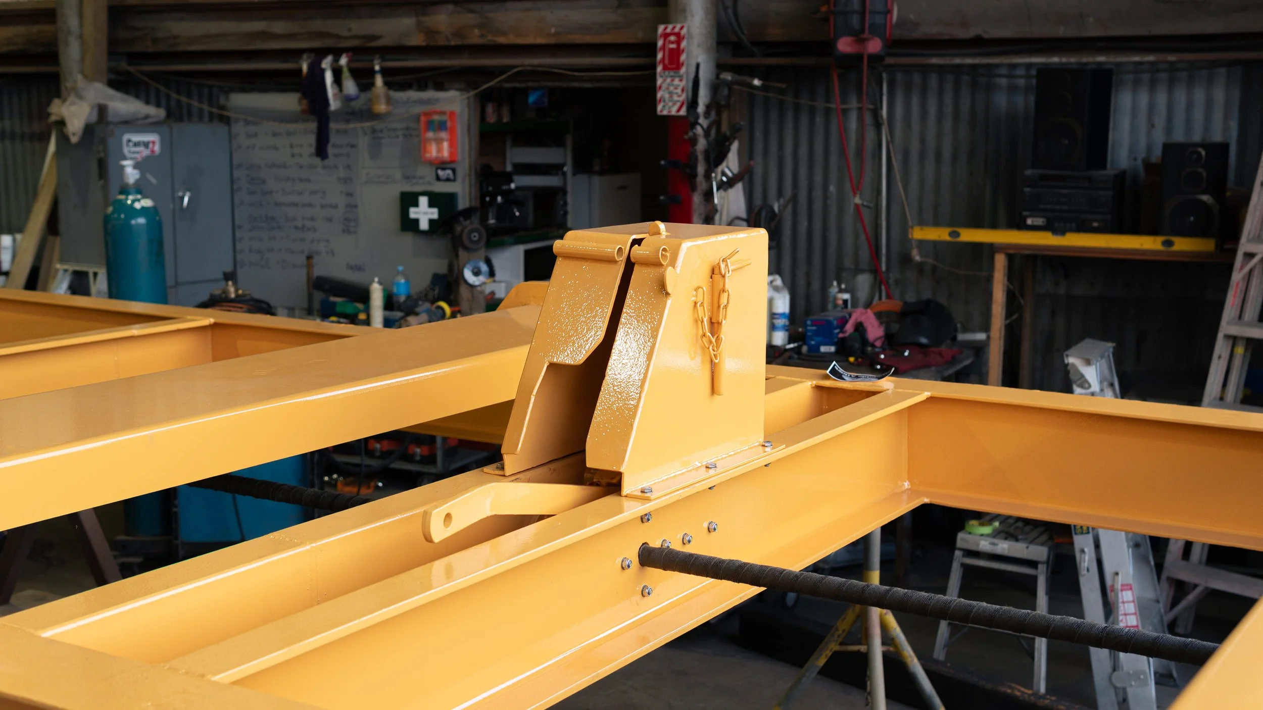 A close-up of yellow metal construction equipment with a hinge, chain, and bolts, inside a workshop with tools, ladders, and shelves in the background.