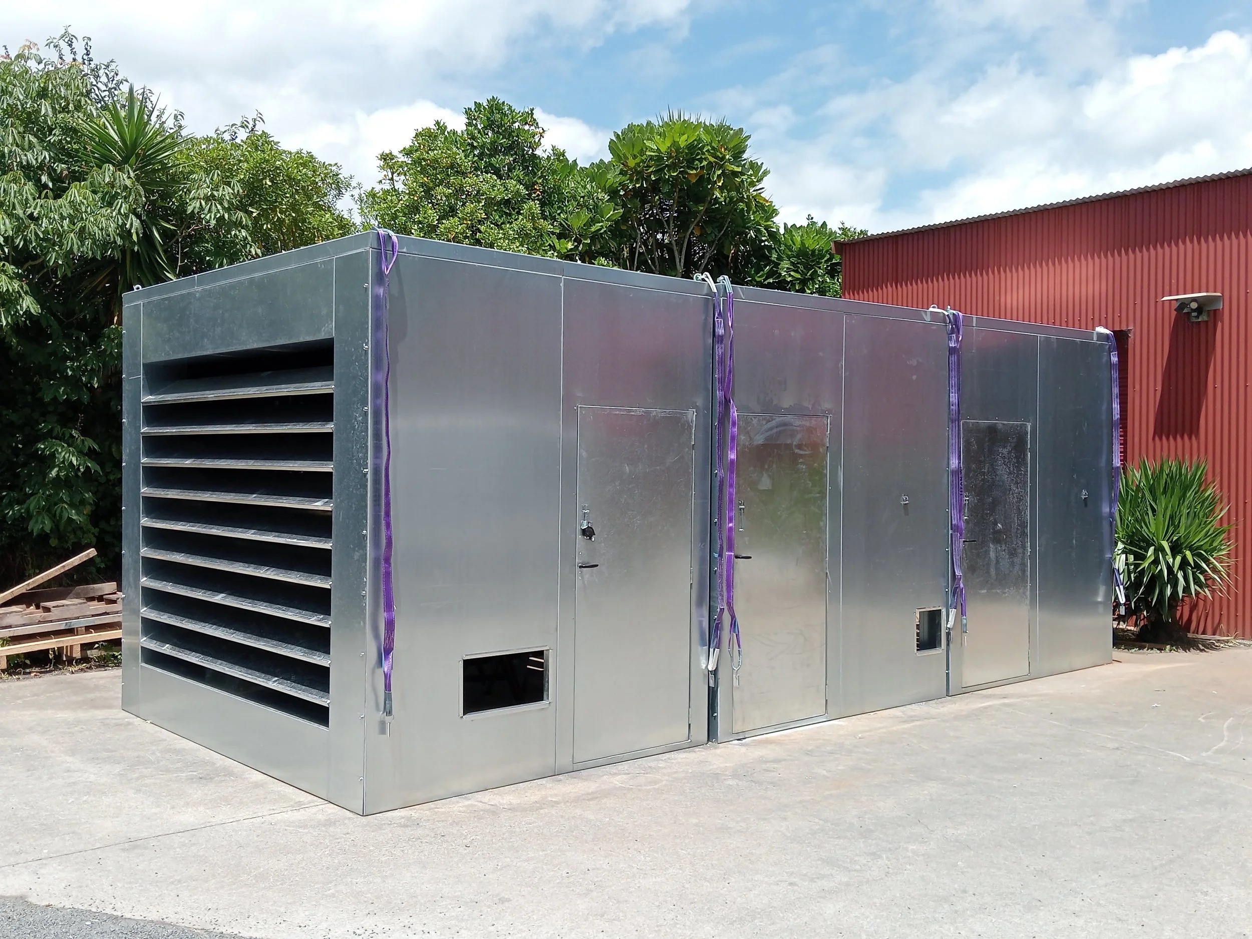Large metal industrial or commercial HVAC unit installed outdoors on concrete ground, with a background of trees and a red building.