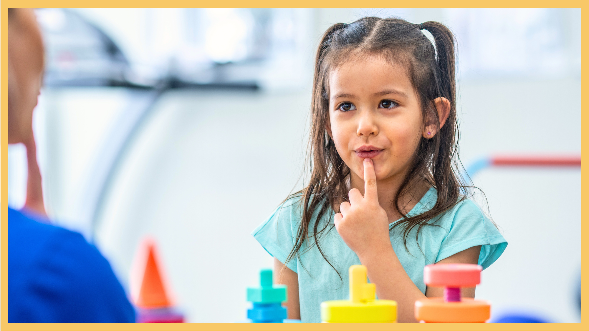 A young girl with long brown hair, wearing a light blue shirt, is sitting at a table with colorful stacking toys. She has her index finger on her lips, practicing lip rounding.