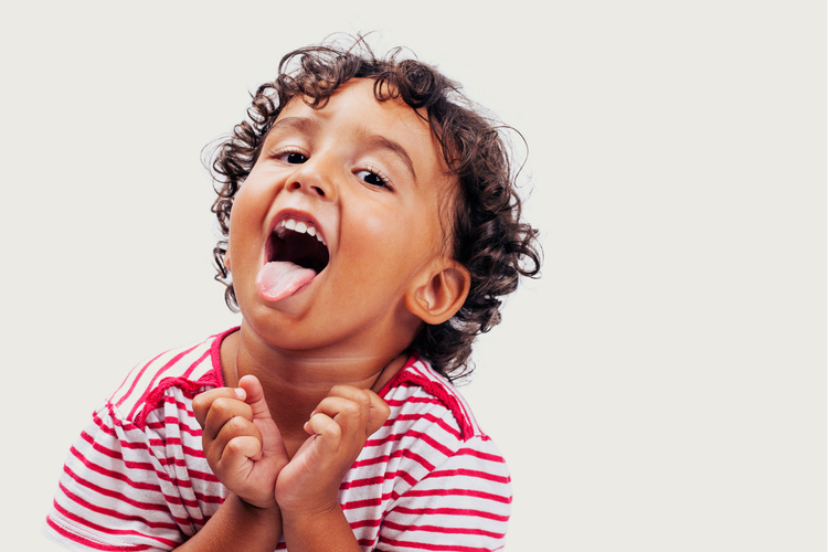 A young girl with long brown hair, wearing a light blue shirt, is sitting at a table with colorful stacking toys. She has her index finger on her lips, practicing lip rounding.